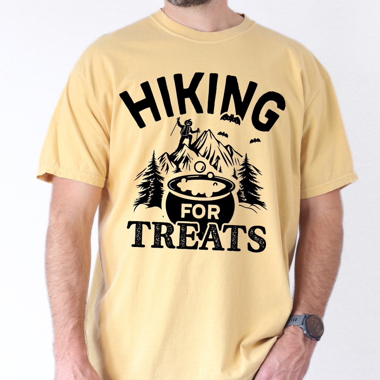 Man wearing Butter Halloween vintage tee Hiking for Treats featuring curved typography bubbling cauldron with candy mountain landscape with character on peak pine trees and flying bats on Comfort Colors shirt for spooky season celebrations for hikers, foodies and adventurers.