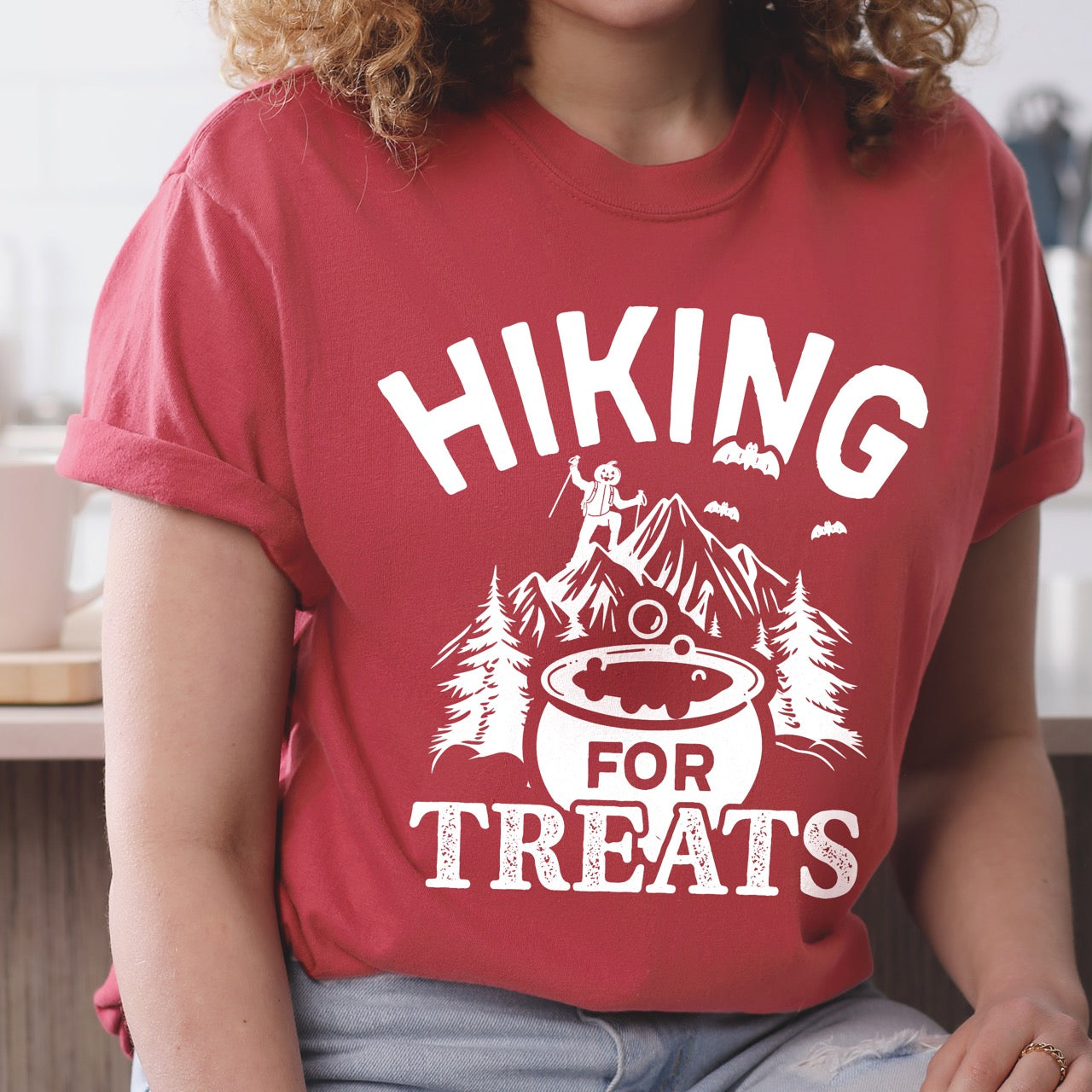 woman wearing crimson Halloween vintage tee Hiking for Treats featuring curved typography bubbling cauldron with candy mountain landscape with character on peak pine trees and flying bats on Comfort Colors shirt for spooky season celebrations for hikers, foodies and adventurers.