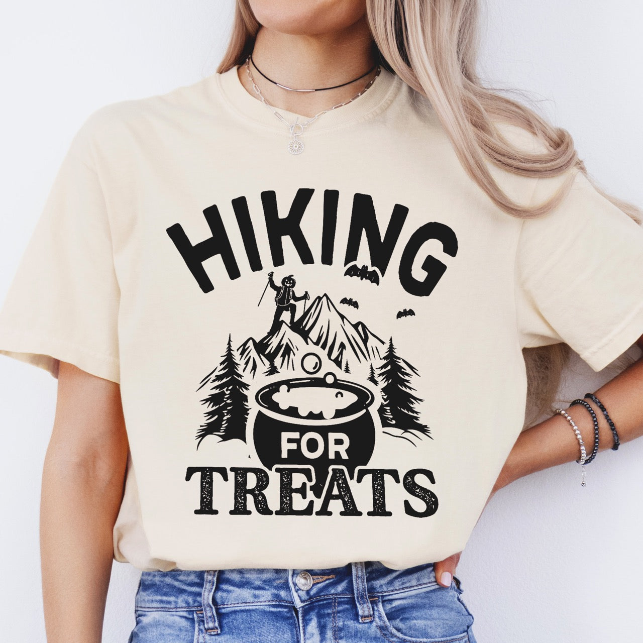 Woman wearing ivory Halloween vintage tee Hiking for Treats featuring curved typography bubbling cauldron with candy mountain landscape with character on peak pine trees and flying bats on Comfort Colors shirt for spooky season celebrations for hikers, foodies and adventurers.
