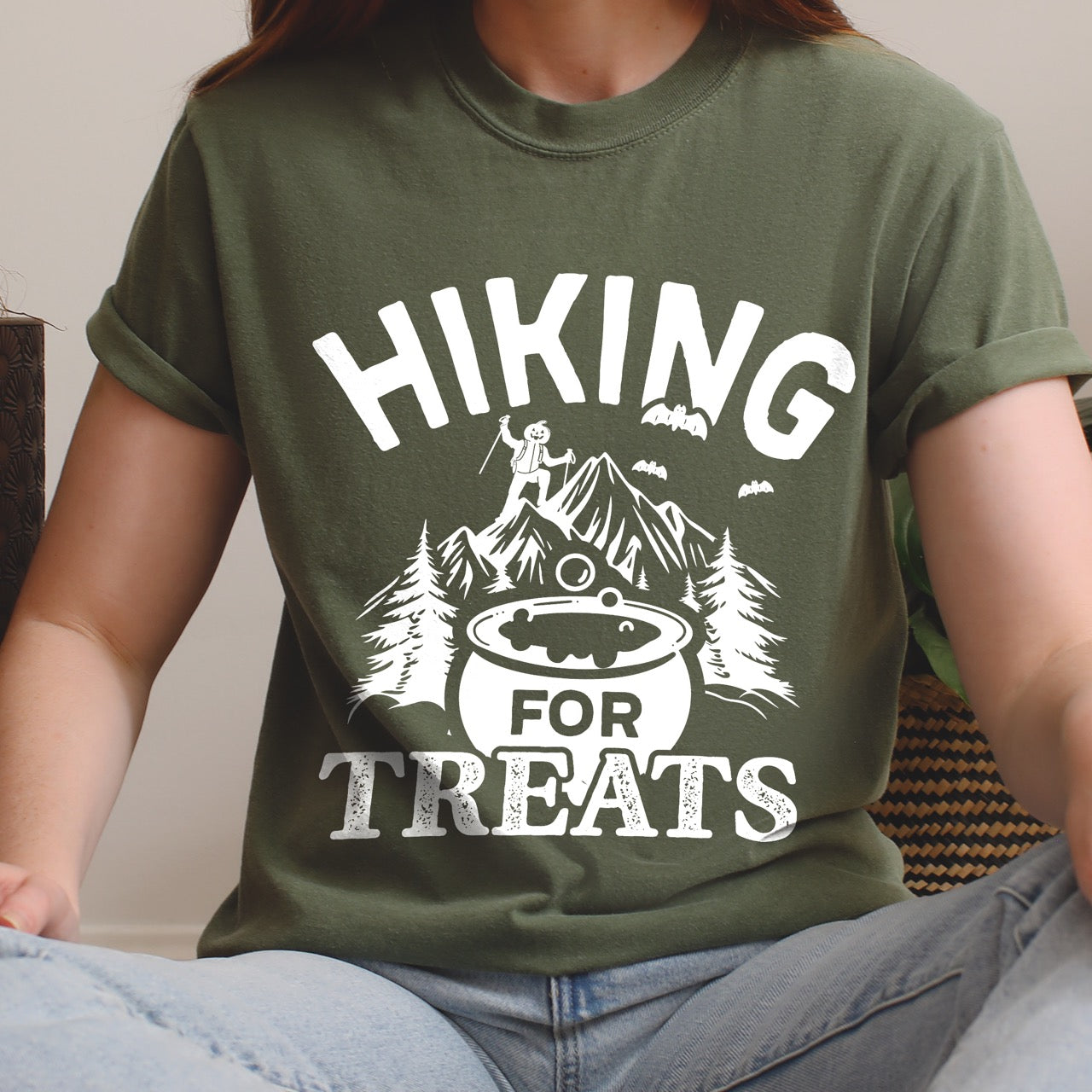 Woman wearing moss Butter Halloween vintage tee Hiking for Treats featuring curved typography bubbling cauldron with candy mountain landscape with character on peak pine trees and flying bats on Comfort Colors shirt for spooky season celebrations for hikers, foodies and adventurers.