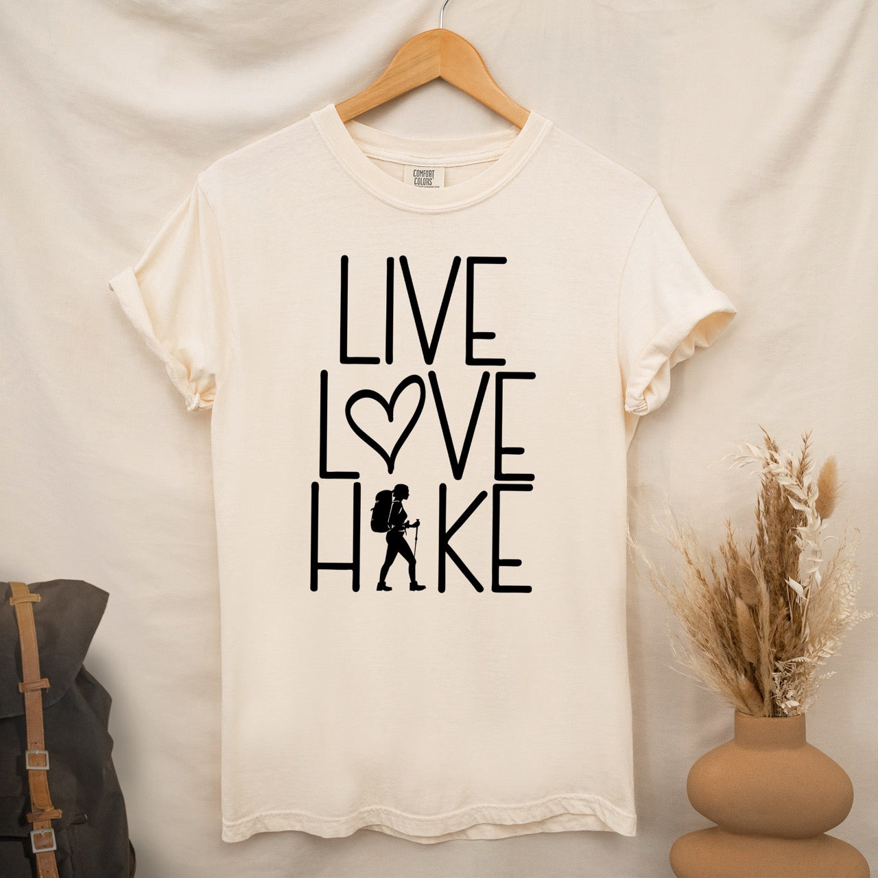 Ivory Women's hiking lifestyle shirt Live Love Hike with minimalist stacked typography heart symbol and female hiker silhouette on Comfort Colors tee for outdoor enthusiasts celebrating trail passion and for hikers and foodies hanging on a wooden hanger
