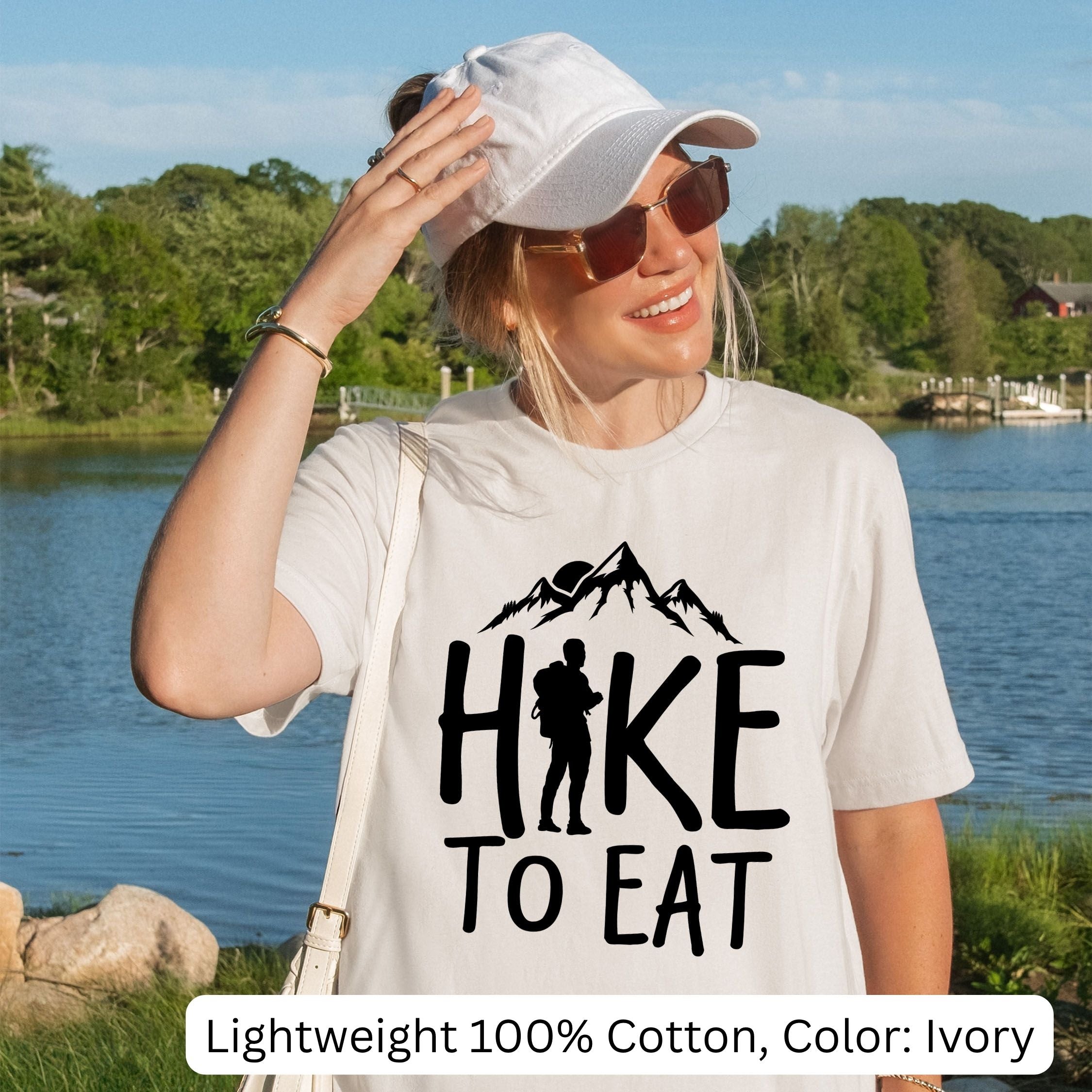 Woman wearing a white t-shirt with 'Hike to Eat' design by a lake.