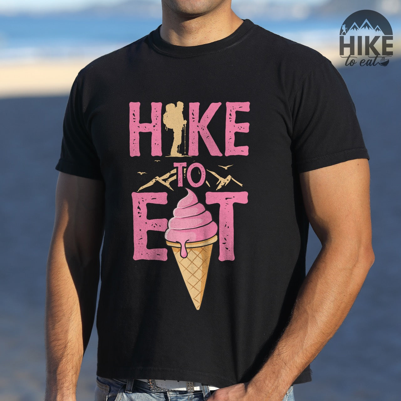 Man wearing black Hike to Eat brand logo t-shirt with distressed pink typography, hiker silhouette integrated into letters, mountain peaks, flying birds, and soft-serve ice cream cone illustration for dessert-loving outdoor enthusiasts