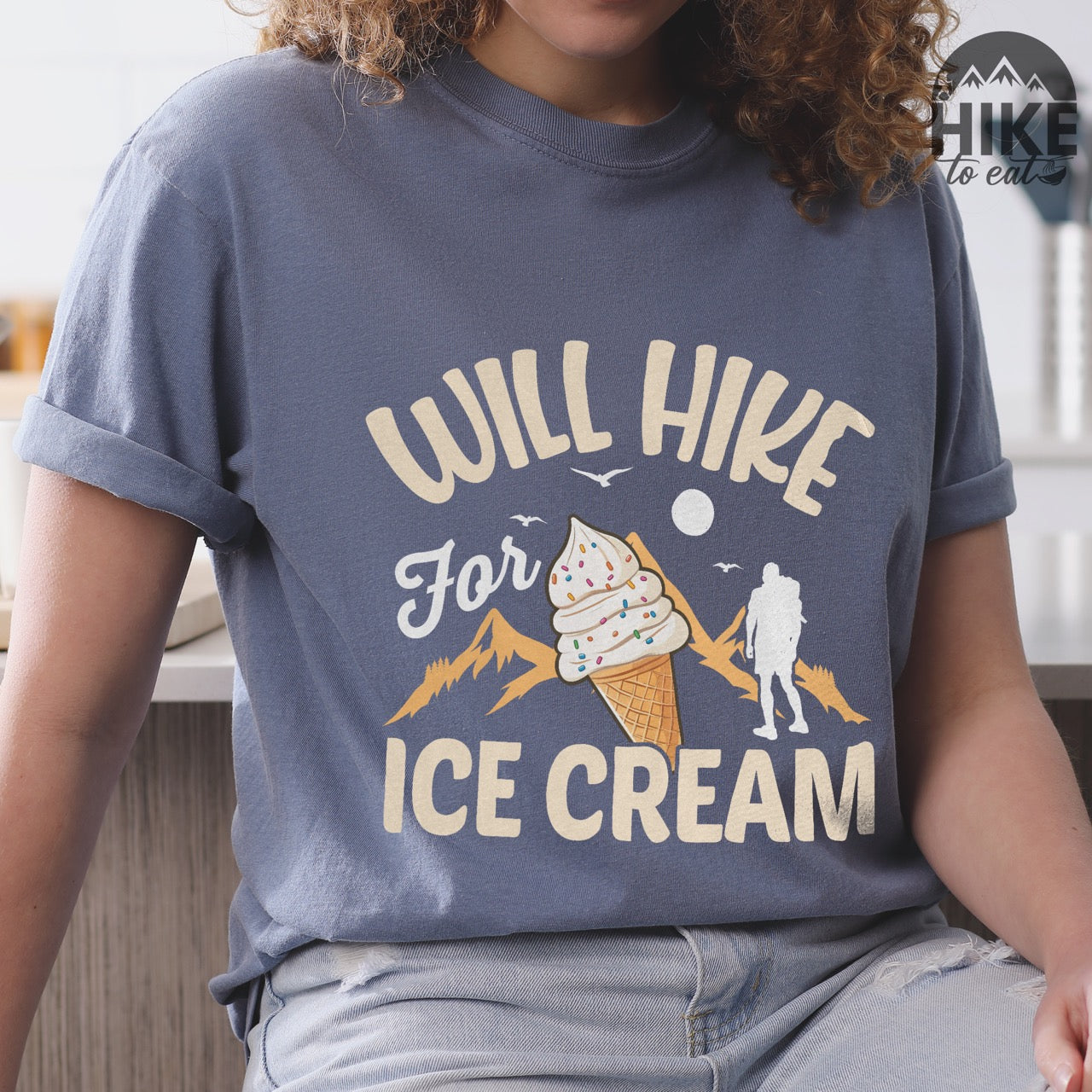 Blue Jean (blue) Will Hike for Ice Cream statement tee with curved typography, script lettering, mountain peaks, hiker silhouette, and ice cream cone illustration on Comfort Colors shirt for dessert-loving outdoor enthusiasts