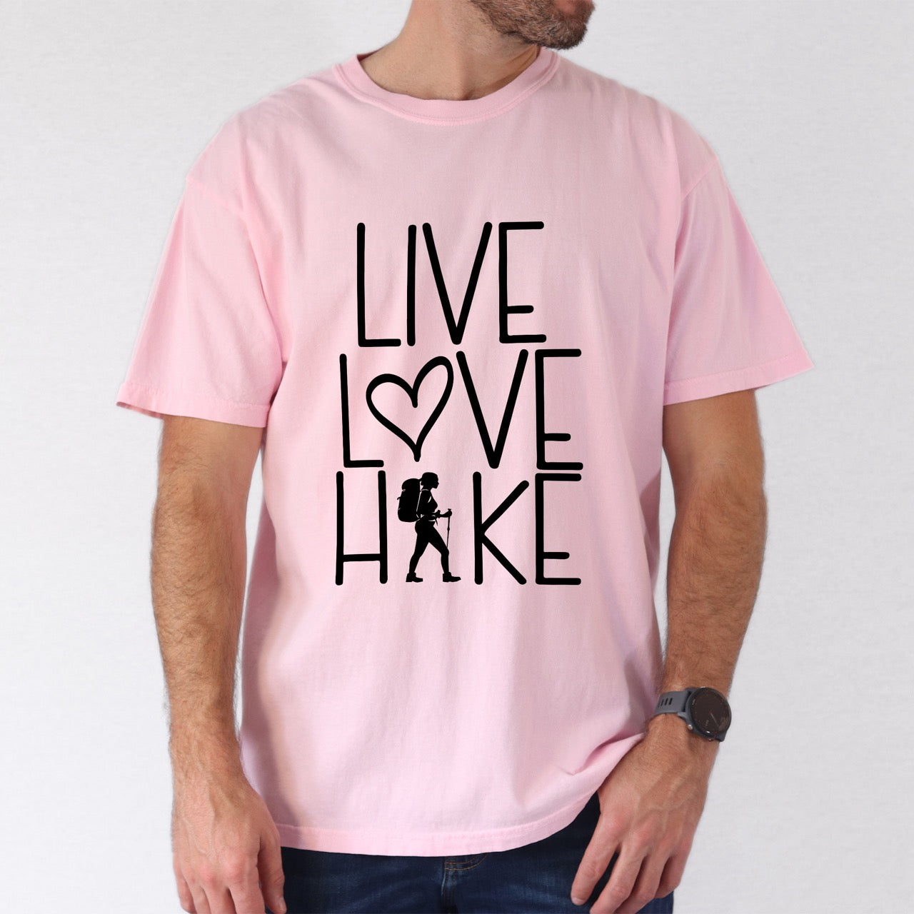 Man wearing Blossom (pink) hiking lifestyle shirt Live Love Hike with minimalist stacked typography heart symbol and female hiker silhouette on Comfort Colors tee for outdoor enthusiasts celebrating trail passion and for hikers and foodies