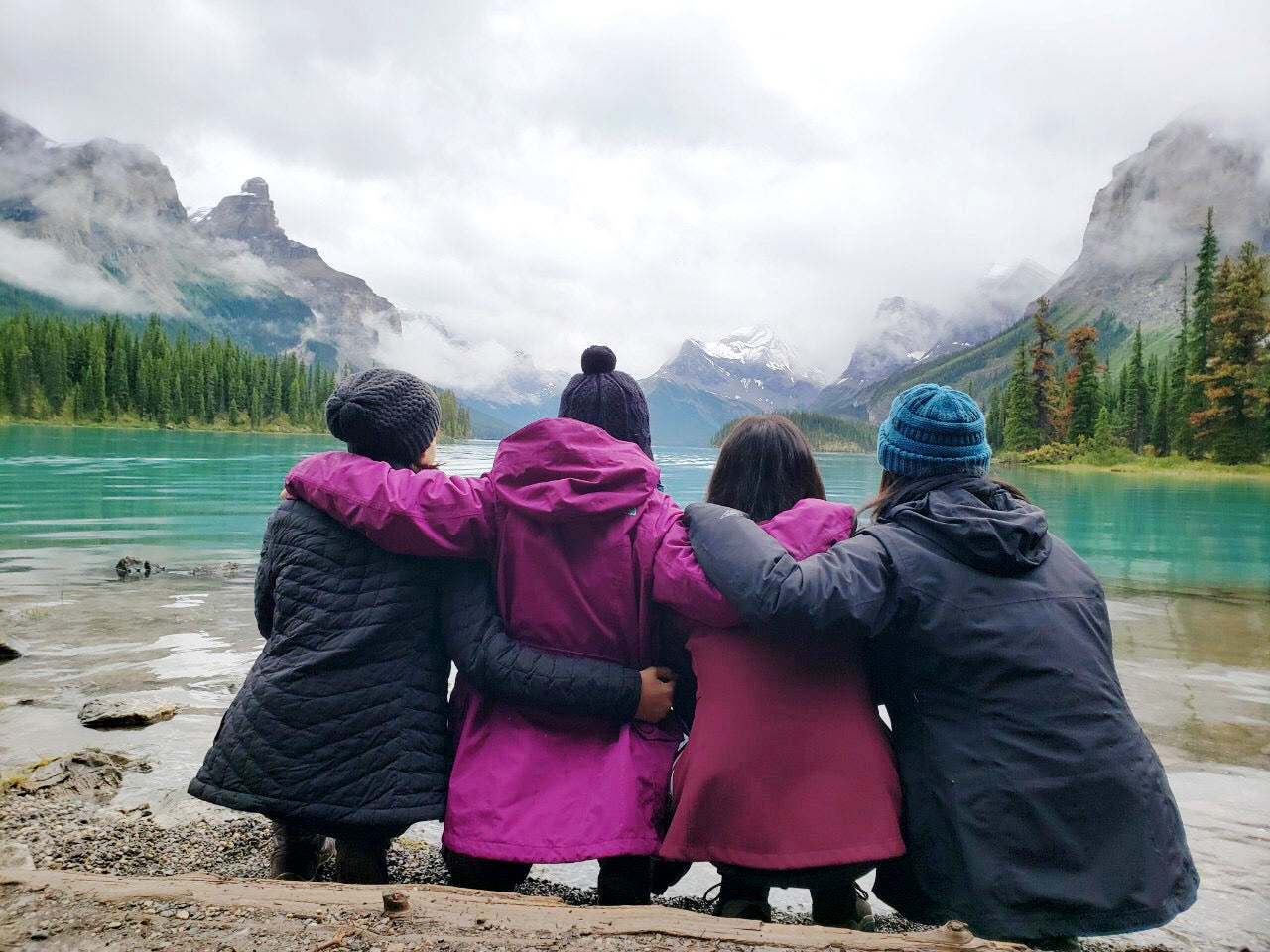 Four people standing with backs to camera at the edge of a turquoise alpine lake. They wear winter jackets (two black, two pink/magenta) and knit hats, with arms around each other's shoulders. They face a landscape of evergreen forest, misty mountains, and cloudy sky. The group stands on a rocky shoreline, viewing the scenic wilderness together.