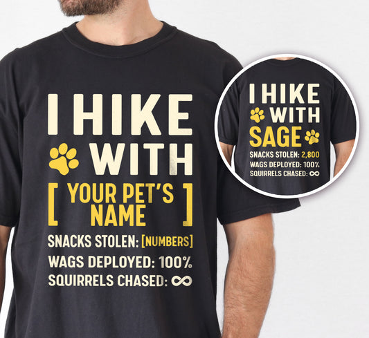 Black graphic t-shirt with 'Personalized Trail Companion' design featuring 'I Hike with' text and customizable pet name field in cream and yellow lettering with paw prints, plus hiking stats with customizable snacks stolen number, wags deployed 100%, and squirrels chased infinity, shown with close-up detail.