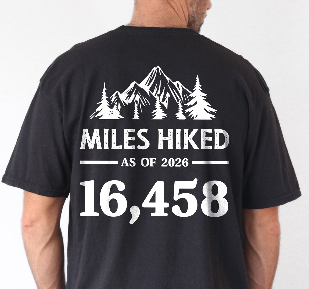 Man wearing black t-shirt with Hike to Eat logo on front chest, back featuring MILES HIKED AS OF 2026 text with personalized number field, mountain and pine tree silhouettes, customizable achievement tracker design - perfect for hikers. 