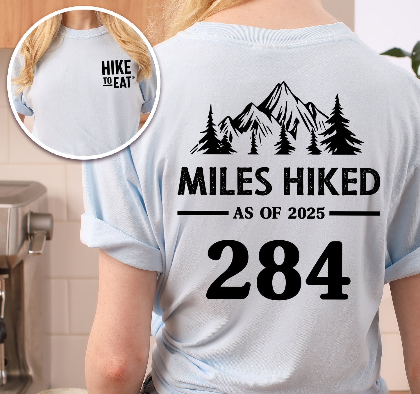 Chambray (Light Blue) Hike to Eat t-shirt front and back view showing small black chest detail and large back design featuring 'Milestone Tracker Personalized' with mountain and pine tree silhouettes, 'Miles Hiked as of 2025' text and customizable numbers field, displaying example number 284.