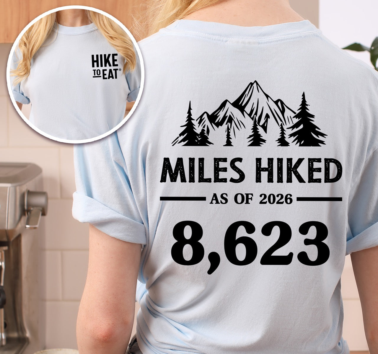 Woman wearing Chambray (light blue) t-shirt with Hike to Eat logo on front chest, back featuring MILES HIKED AS OF 2026 text with personalized number field, mountain and pine tree silhouettes, customizable achievement tracker design - perfect for hikers. 