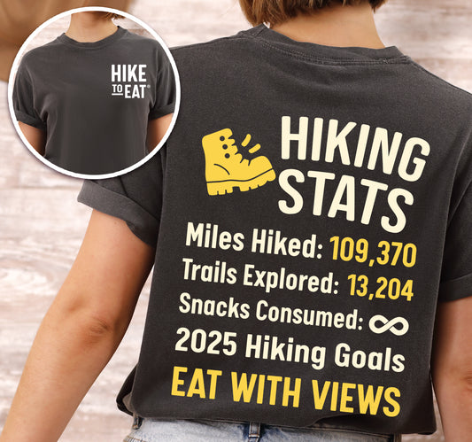 Pepper (Dark Gray) Hike to Eat t-shirt front and back view showing small chest detail and large back design featuring 'Hiking Stats' design with yellow hiking boot icon, customizable miles hiked and trails explored numbers, snacks consumed infinity, and 2025 hiking goals 'Eat with Views'. 