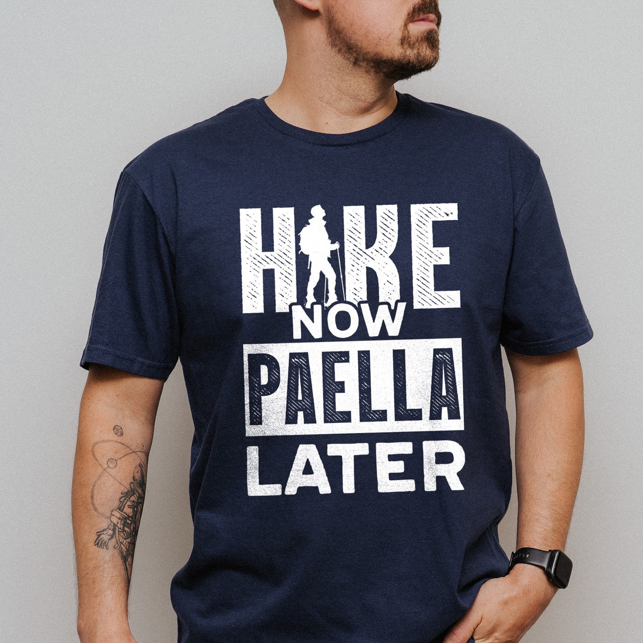 man wearing navy blue Hike Now Paella Later statement tee with bold distressed typography and hiker silhouette for Spanish food-loving outdoor enthusiasts and hikers