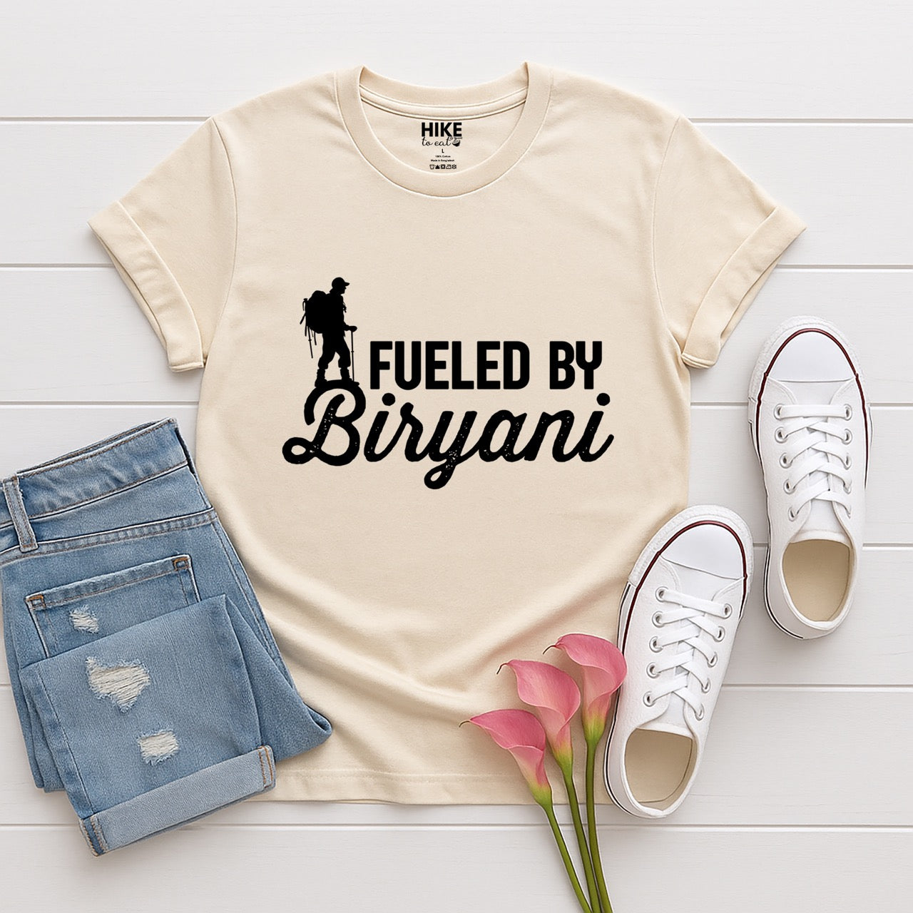 Natural (beige) Fueled by Biryani statement tee with bold typography, elegant script lettering, and hiker silhouette for Indian food-loving outdoor enthusiasts, foodies and hikers with jeans and shoes