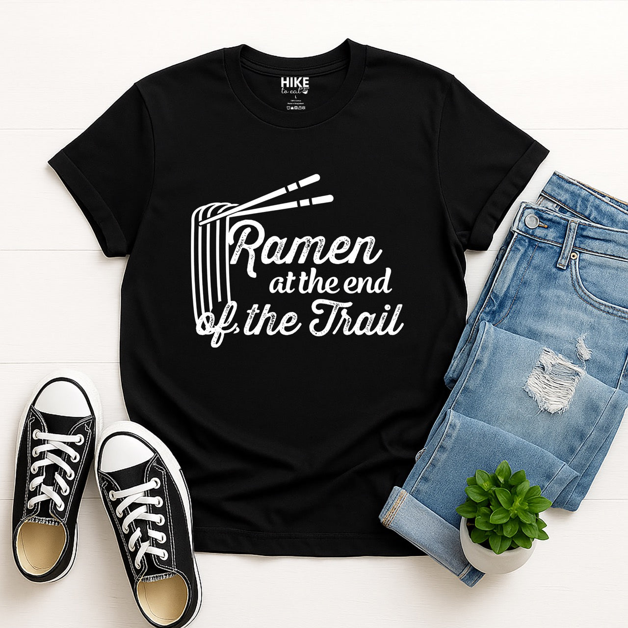 Black Ramen at the End of the Trail statement tee with elegant script typography, ramen bowl illustration, and chopsticks for Japanese noodle-loving outdoor enthusiasts, foodies and hikers flat layed with jeans and shoes