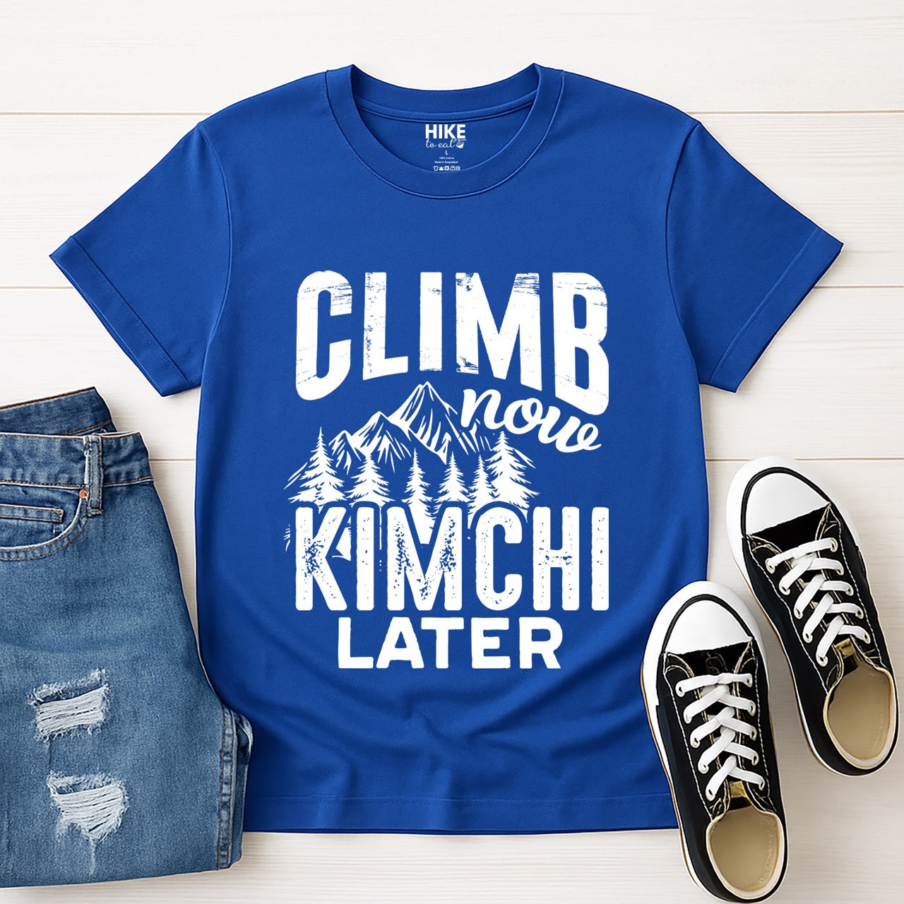 Royal blue Climb now Kimchi later statement tee with distressed bold typography, script lettering, mountain peaks, and pine trees for Korean food-loving climbers, hikers and foodies laid flat with jeans and shoes