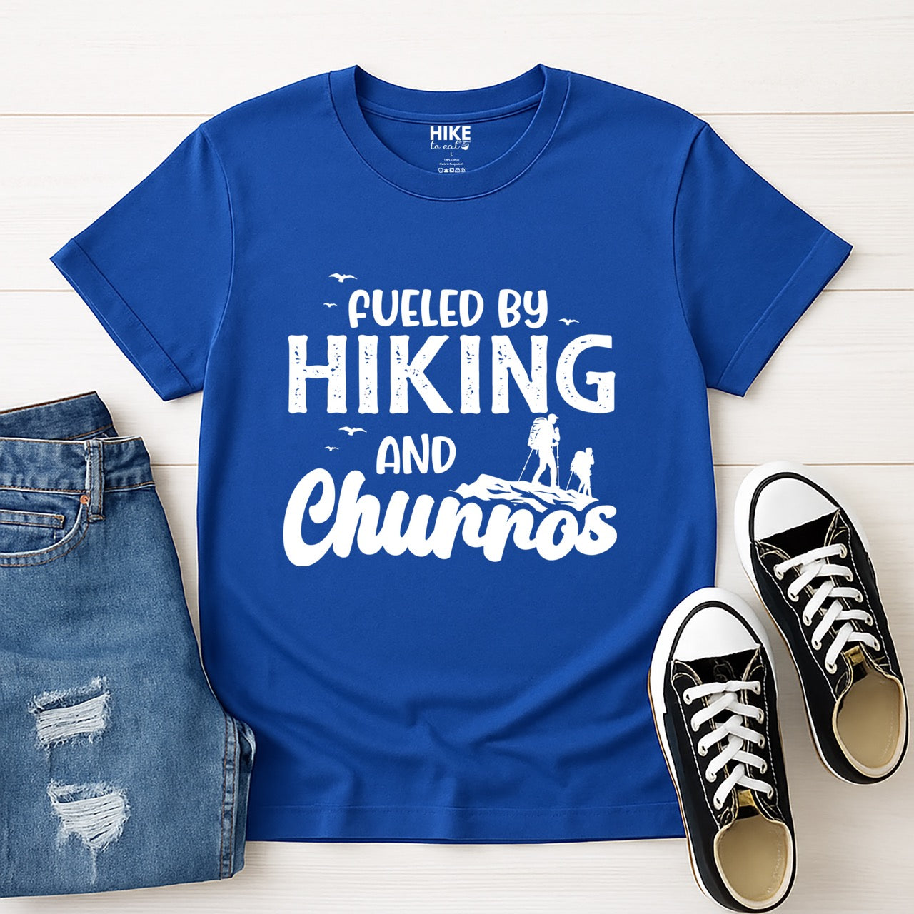 Royal blue Fueled by Hiking and Churros statement tee with bold distressed typography, flying birds, and two hiker silhouettes on mountain ridge for Spanish dessert-loving outdoor enthusiasts, hikers and foodies laid flat with shoes and pants