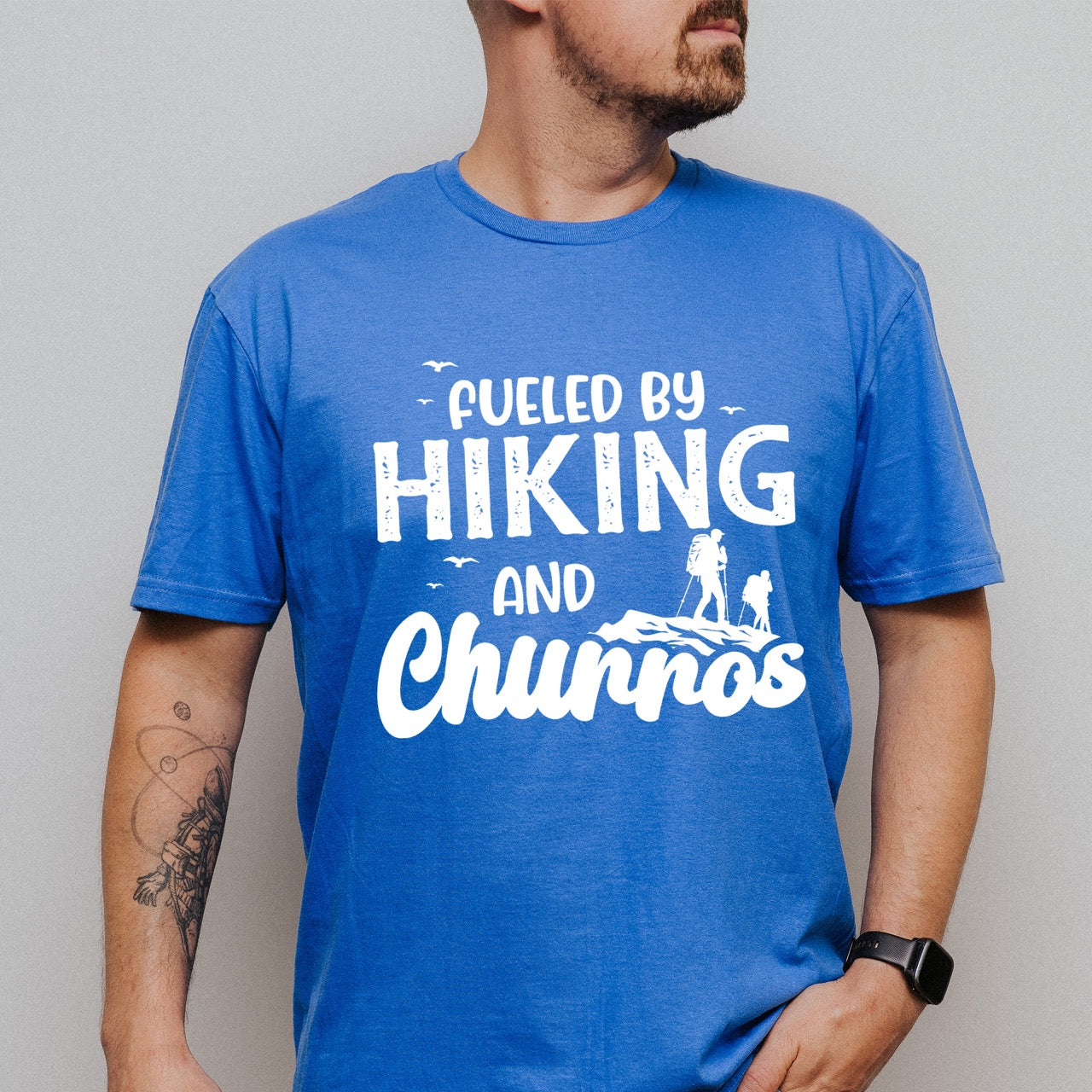Man wearing Royal blue Fueled by Hiking and Churros statement tee with bold distressed typography, flying birds, and two hiker silhouettes on mountain ridge for Spanish dessert-loving outdoor enthusiasts, hikers and foodies
