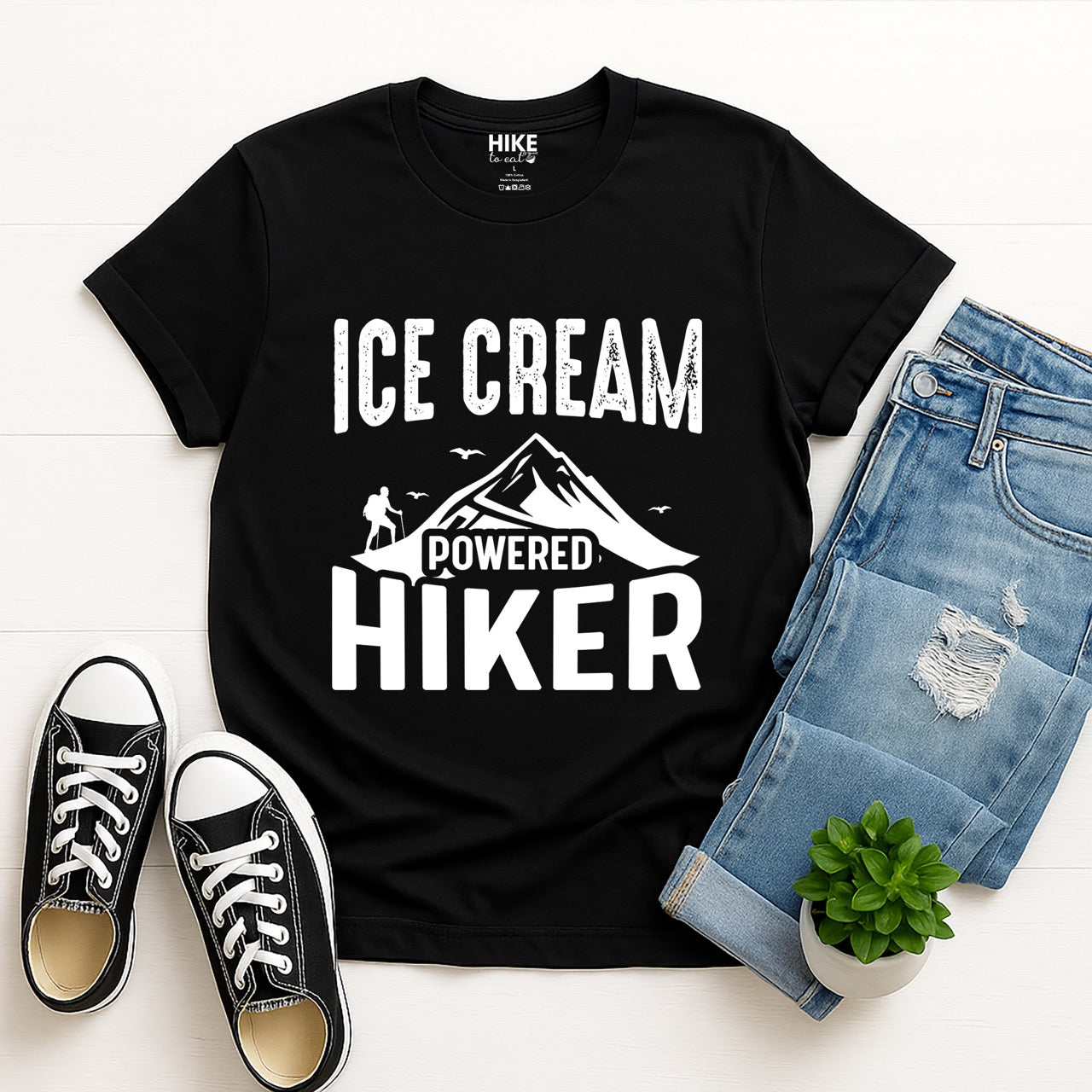 Black  Ice Cream Powered Hiker statement tee with bold distressed typography, mountain peaks, hiker silhouette, and flying birds for dessert-loving outdoor enthusiasts, hikers and foodies laid flat with jeans and shoes