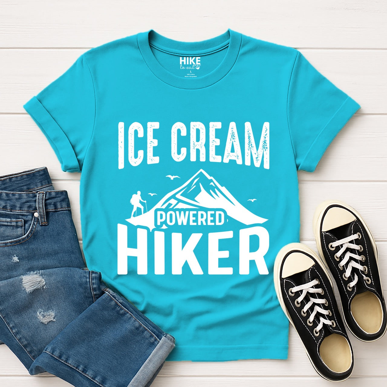 Tropical blue Ice Cream Powered Hiker statement tee with bold distressed typography, mountain peaks, hiker silhouette, and flying birds for dessert-loving outdoor enthusiasts, hikers and foodies laid flat with shoes and jeans