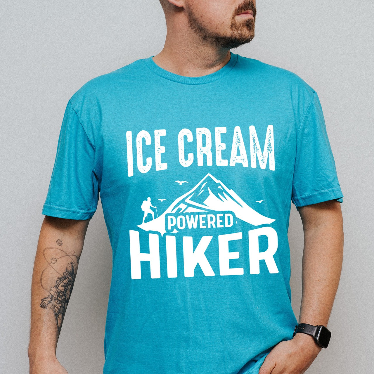 Man wearing Tropical blue Ice Cream Powered Hiker statement tee with bold distressed typography, mountain peaks, hiker silhouette, and flying birds for dessert-loving outdoor enthusiasts, hikers and foodies