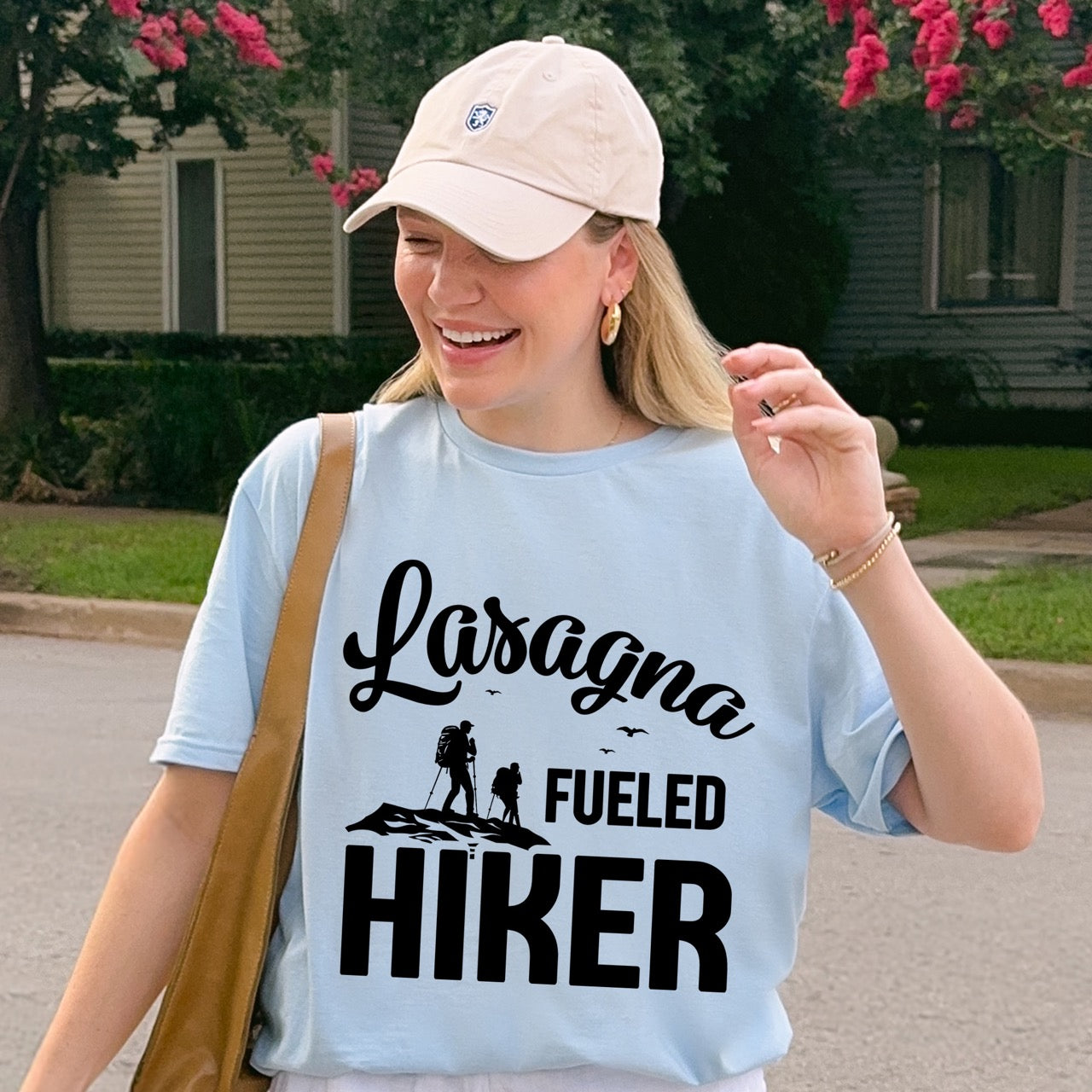 Woman wearing Light Blue Lasagna Fueled Hiker statement tee with flowing script typography, bold lettering, two hiker silhouettes on mountain ridge, and flying birds for Italian food-loving outdoor enthusiasts, hikers and foodies