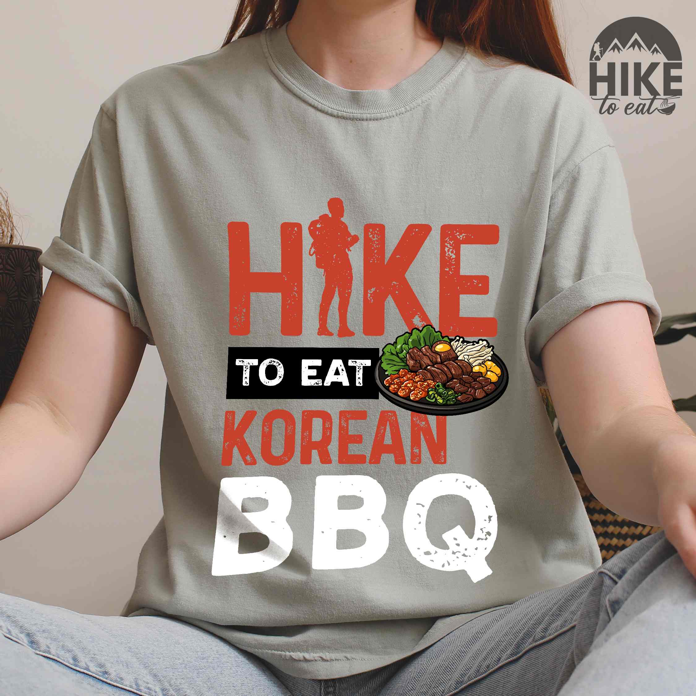 woman wearing bay "Hike to eat Korean bbq" colored vintage tee with bold typography, illustrated grilling platter korean bbq, mountains, birds, and backpacker for kbbq lovers.