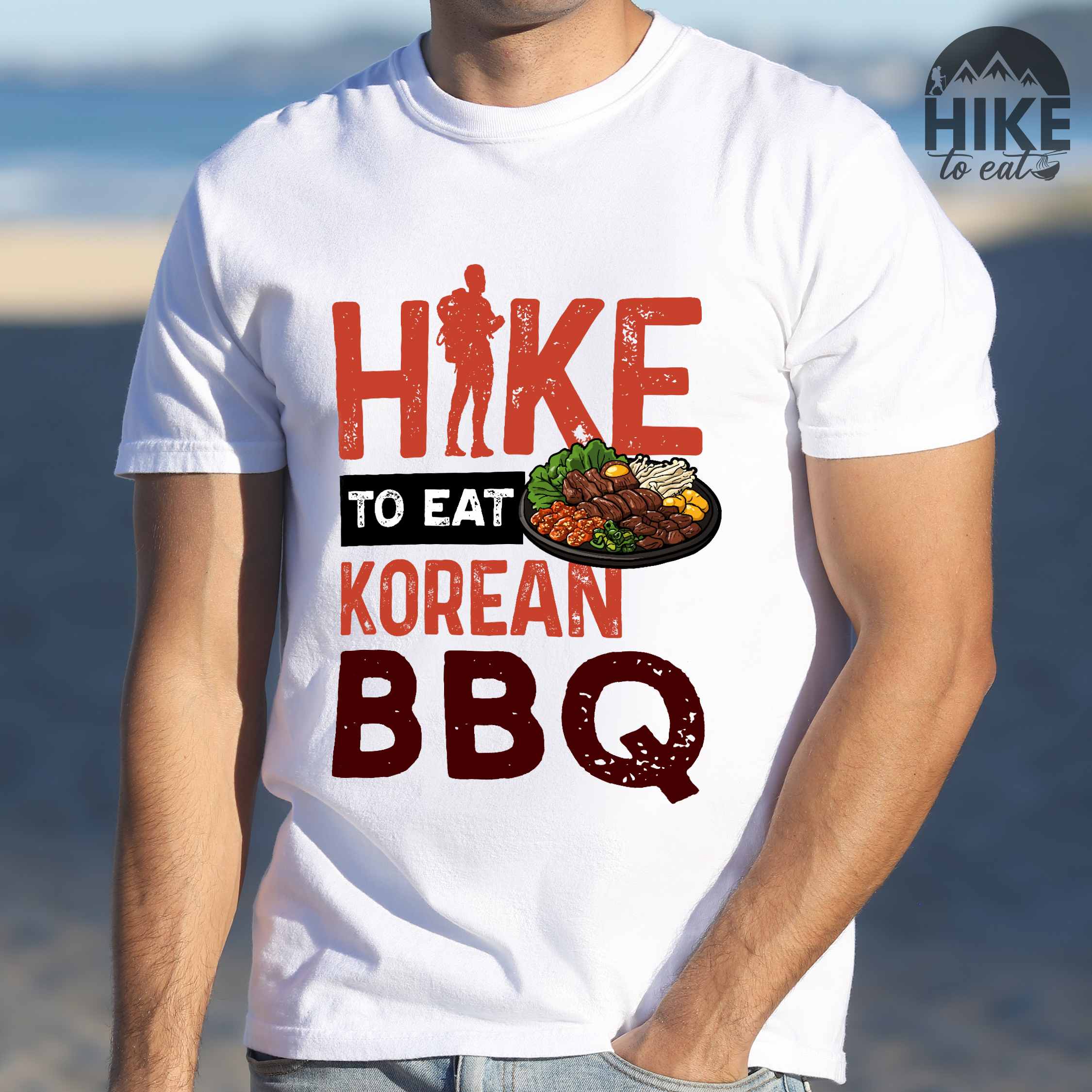 Man wearing white "Hike to eat Korean bbq" colored vintage tee with bold typography, illustrated grilling platter korean bbq, mountains, birds, and backpacker for kbbq lovers.