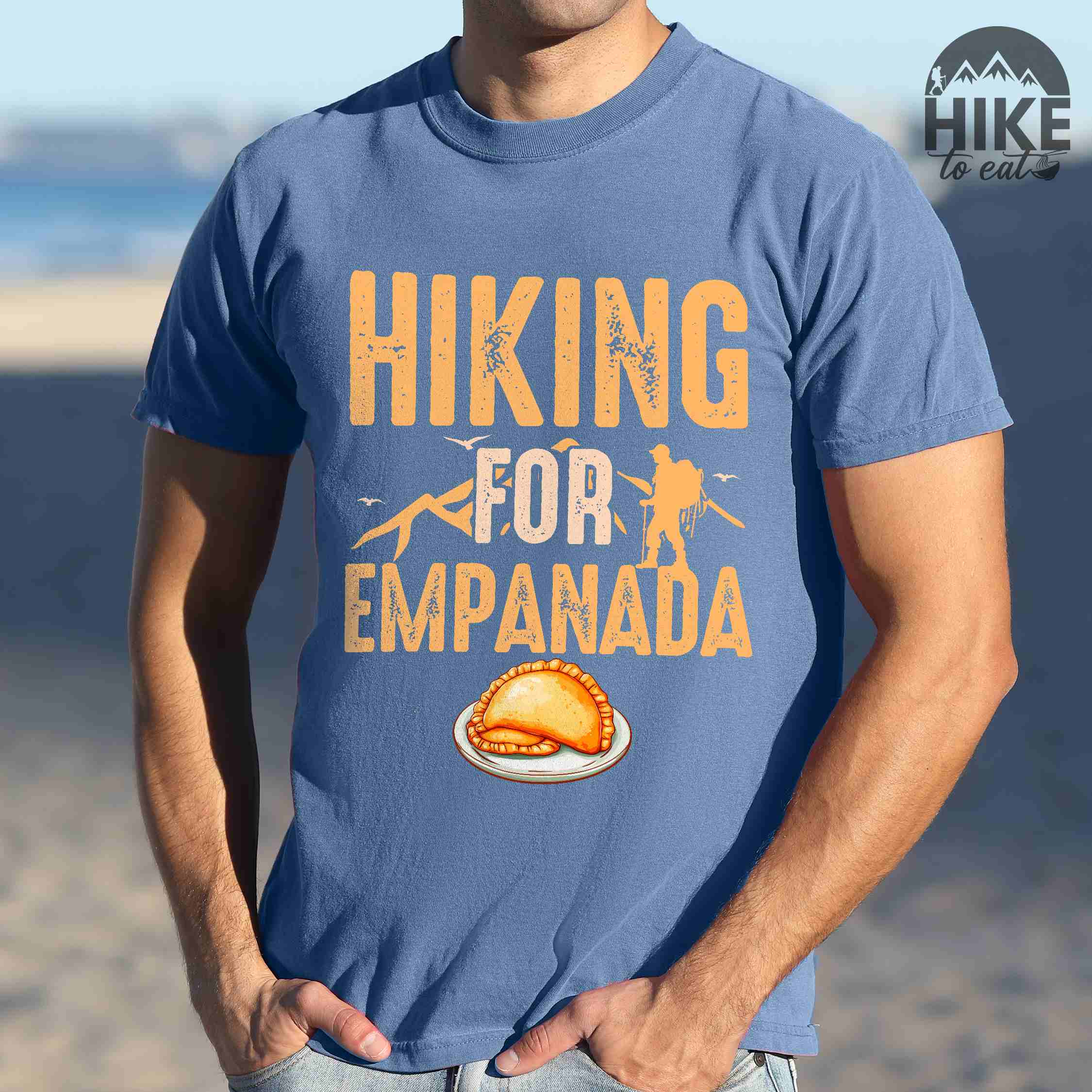Man wearing blue jean "Hiking for empanada" colored vintage tee with bold typography, illustrated freshly baked empanada, mountains, birds, and backpackers for asian cuisine enthusiasts.