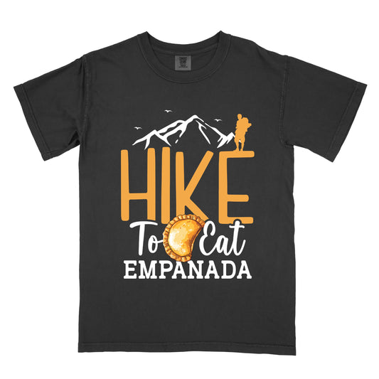 Black t-shirt with golden orange and white text reading "HIKE To Eat EMPANADA". "HIKE" appears in large golden orange letters in the middle, "To Eat" in white script below it, and "EMPANADA" in white letters at the bottom. Features white mountain outlines with small white birds flying above, a golden orange silhouette of a hiker at the top right of the mountains, and an illustration of an empanada between "To" and "Eat". 