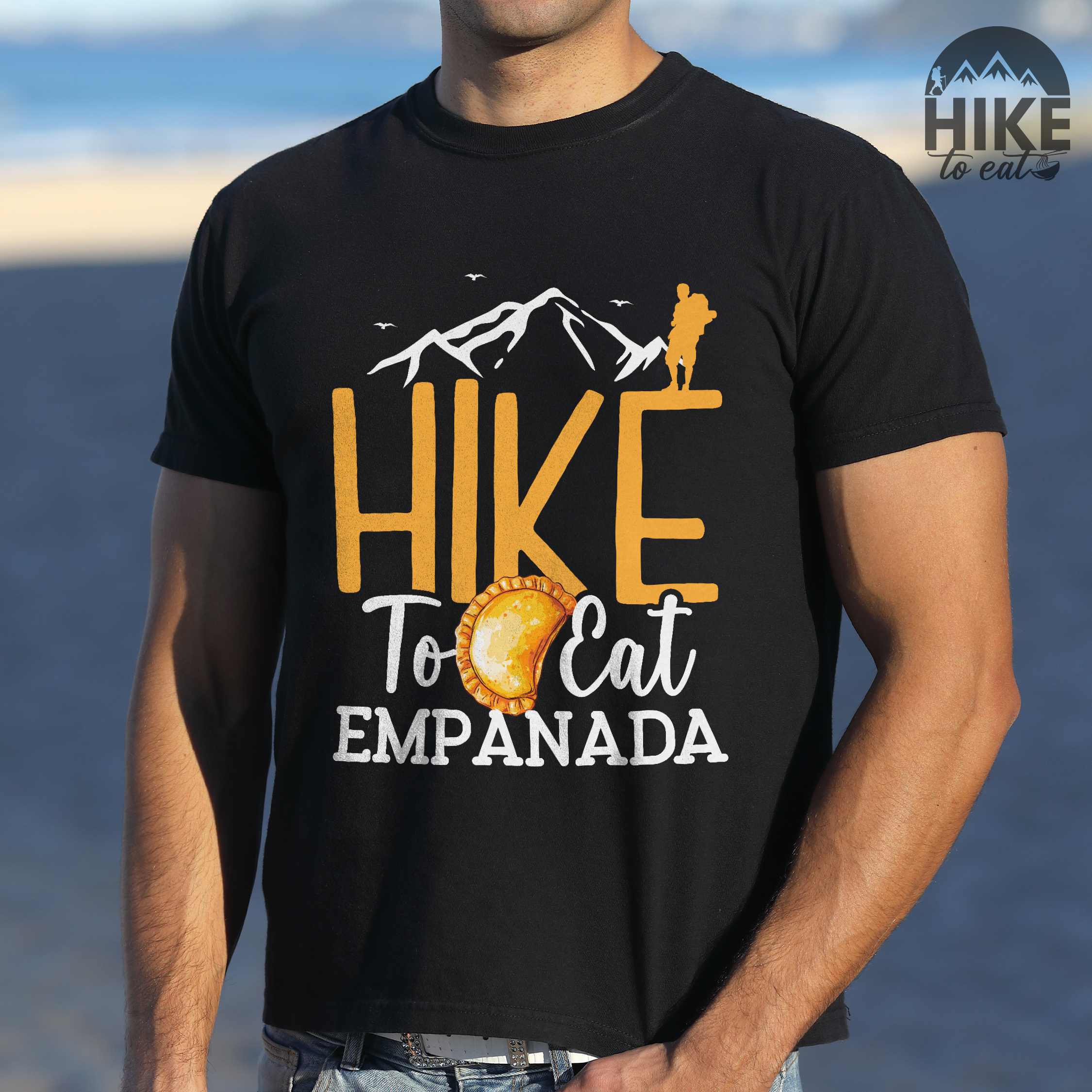 Man wearing black "Hike to eat empanada" colored vintage tee with bold typography, illustrated freshly baked empanada, mountains, birds, and backpackers for street food enthusiasts.