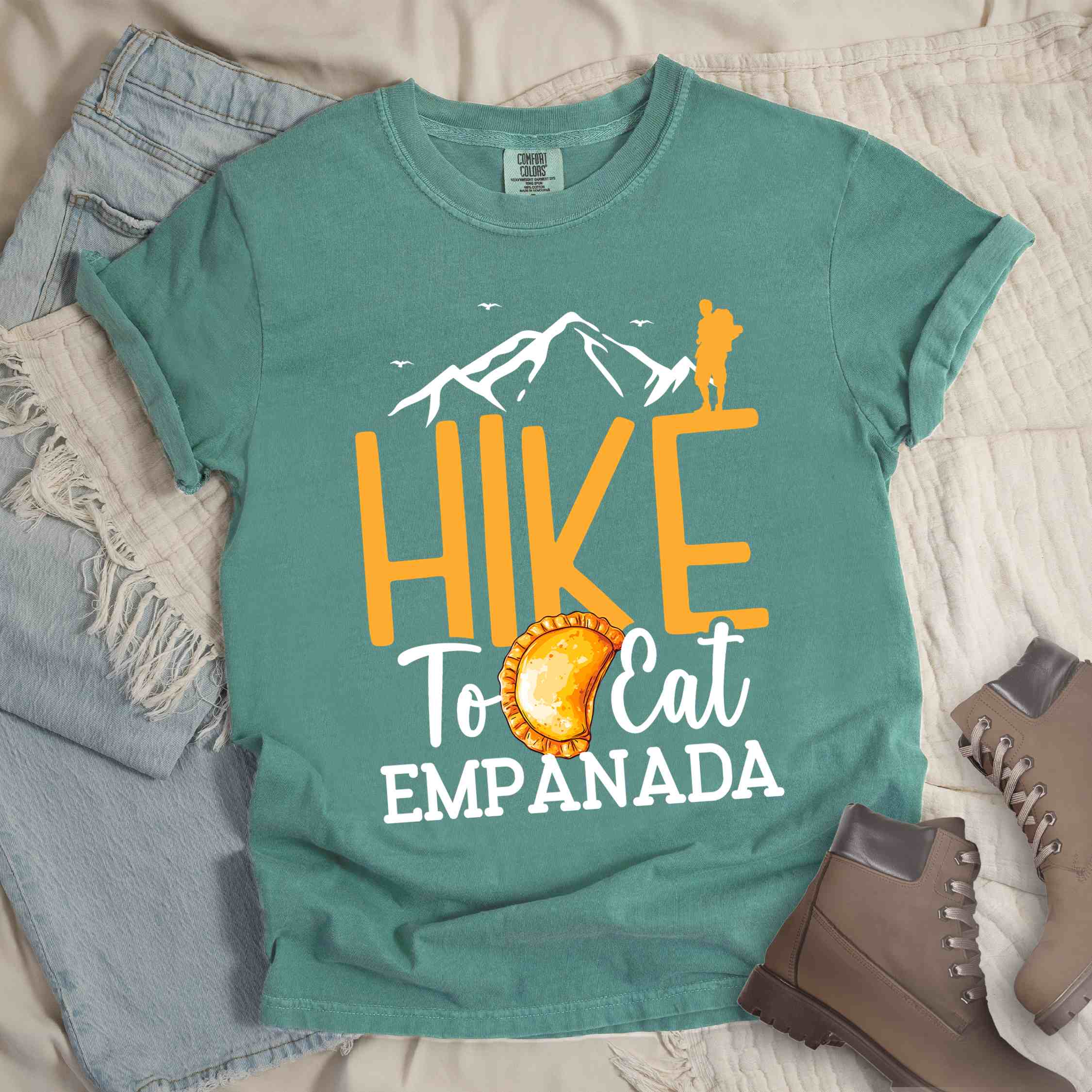 Light green "Hike to eat empanada" colored vintage tee with bold typography, illustrated freshly baked empanada, mountains, birds, and backpackers for street food enthusiasts.