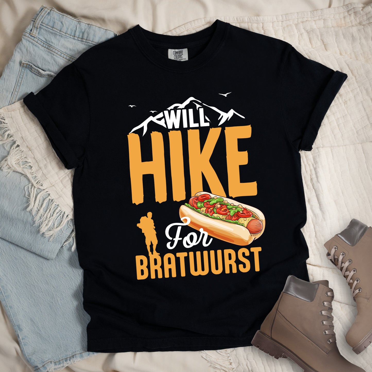 Black t-shirt with white, golden orange, and white text reading "WILL HIKE For BRATWURST". "WILL" appears in white text integrated with mountain outlines at the top, "HIKE" in large golden orange letters in the middle, "For" in white script below it, and "BRATWURST" in golden orange at the bottom. Features an illustration of a bratwurst in a bun with toppings to the right of "For" and a golden orange silhouette of a hiker to the left of "BRATWURST". Small white birds fly near the mountains. 
