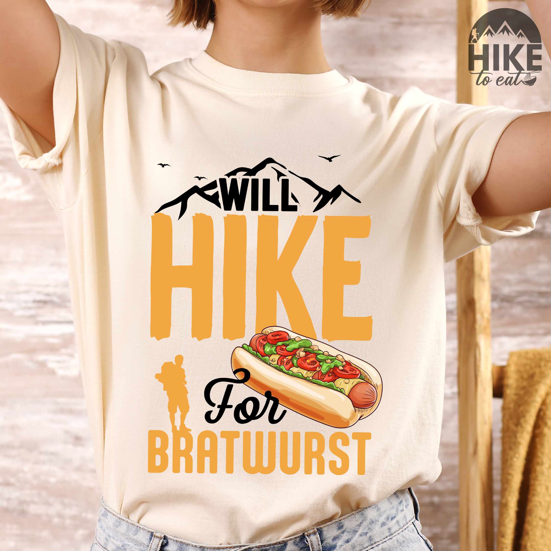Woman wearing ivory "Will hike for bratwurst" colored vintage tee with bold typography, illustrated bratwurst in a bun, mountains, birds, and backpackers for street food enthusiasts.
