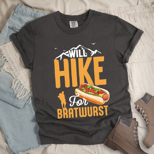 Pepper (Dark Gray) t-shirt with white, golden orange, and white text reading "WILL HIKE For BRATWURST". "WILL" appears in white text integrated with mountain outlines at the top, "HIKE" in large golden orange letters in the middle, "For" in white script below it, and "BRATWURST" in golden orange at the bottom. Features an illustration of a bratwurst in a bun with toppings to the right of "For" and a golden orange silhouette of a hiker to the left of "BRATWURST". Small white birds fly near the mountains.