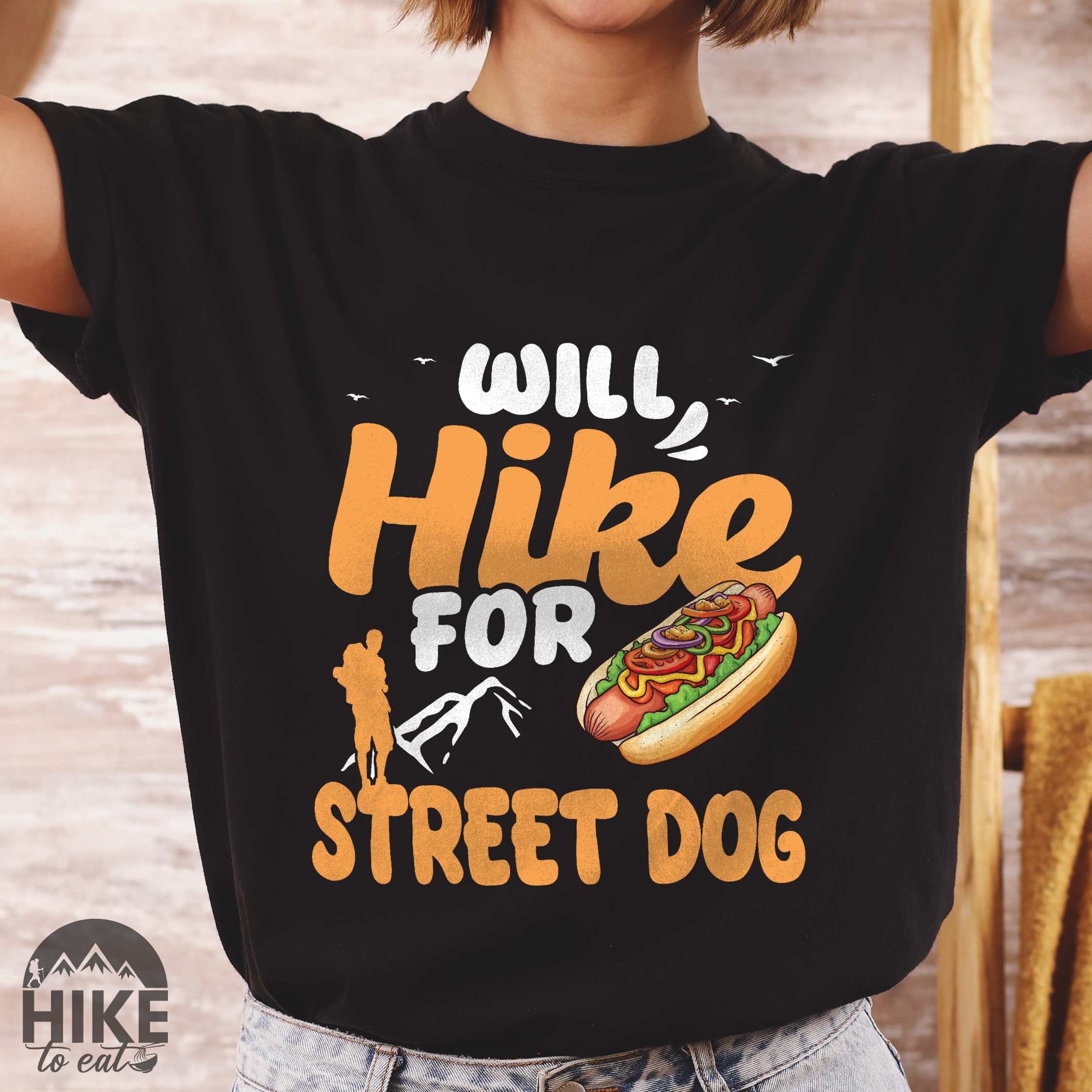 Woman wearing black "Will hike for street dog" colored vintage tee with bold typography, illustrated freshly bratwurst in a bun, mountains, birds, and backpackers for street dog lovers.