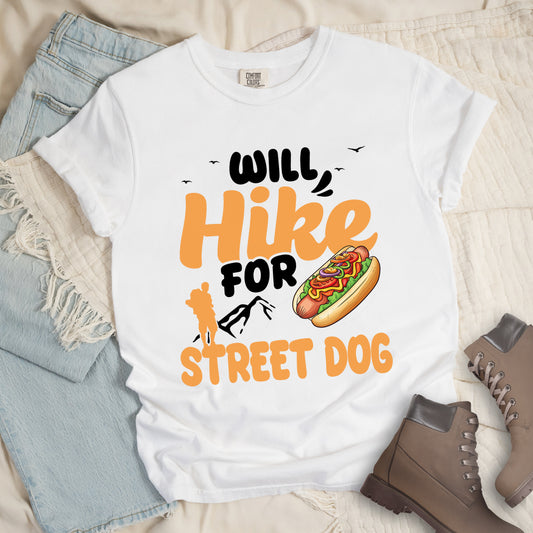 White t-shirt with orange and black text reading "WILL Hike FOR STREET DOG". "WILL" appears in black at the top with small black birds flying nearby, "Hike" in large orange script in the middle, "FOR" in black below it with small black mountain outlines, and "STREET DOG" in orange at the bottom. Features an illustration of a hot dog with toppings to the right of "FOR" and an orange silhouette of a hiker to the left top of "STREET DOG".