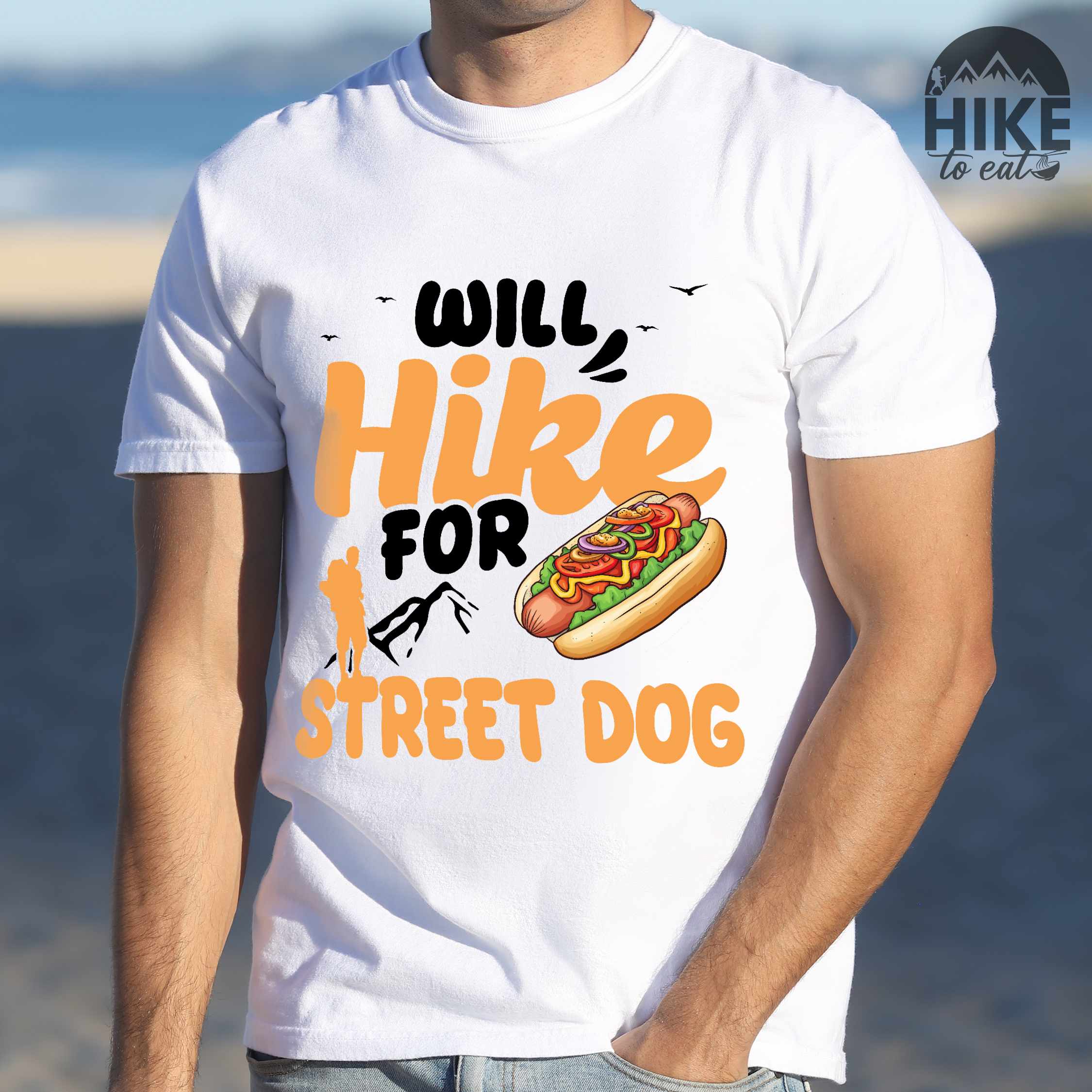 Man wearing white "Will hike for street dog" colored vintage tee with bold typography, illustrated freshly bratwurst in a bun, mountains, birds, and backpackers for street dog lovers.