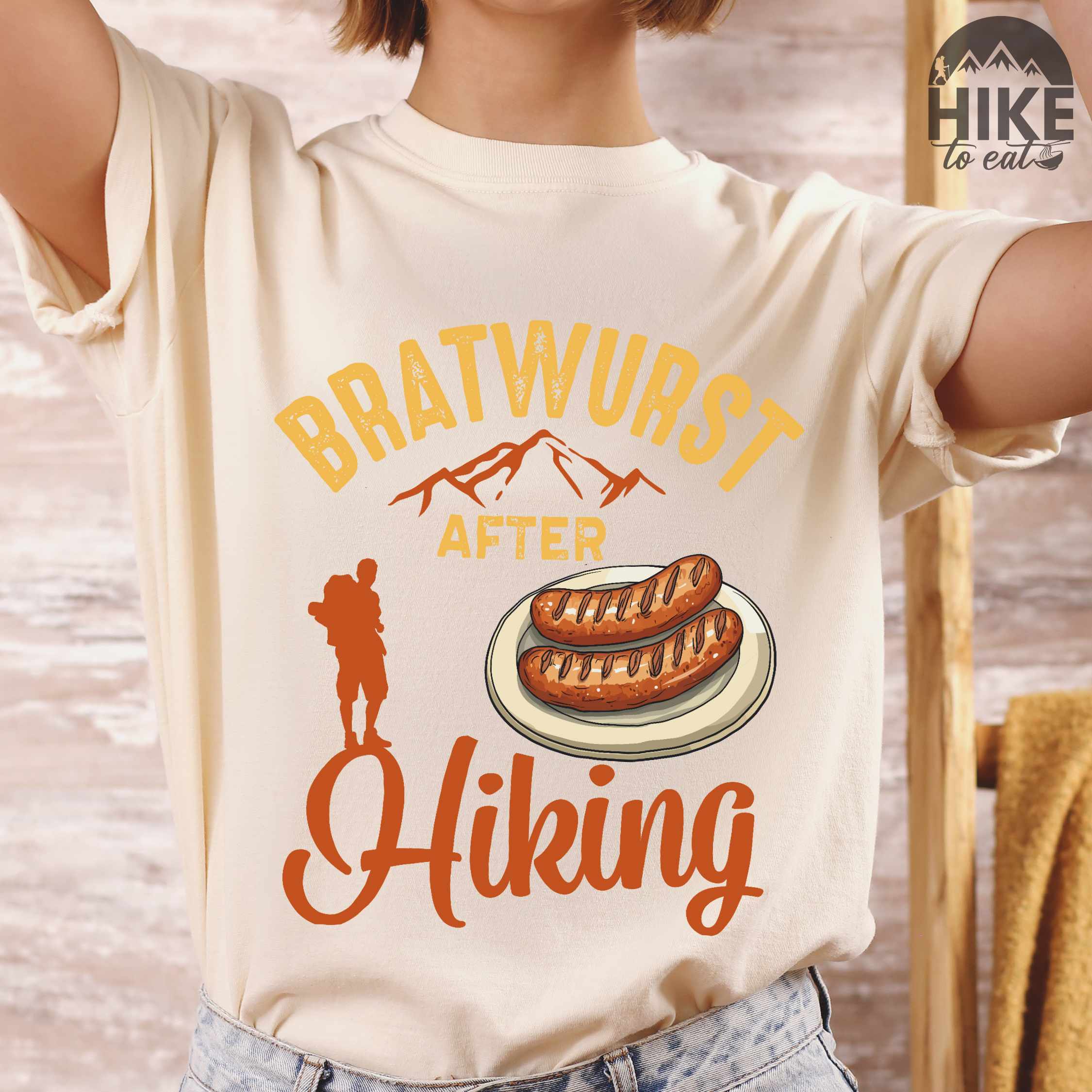woman wearing ivory "bratwurst after hiking" colored vintage tee with bold typography, illustrated bratwurst on a plate, mountains, birds, and backpackers for street food enthusiasts.