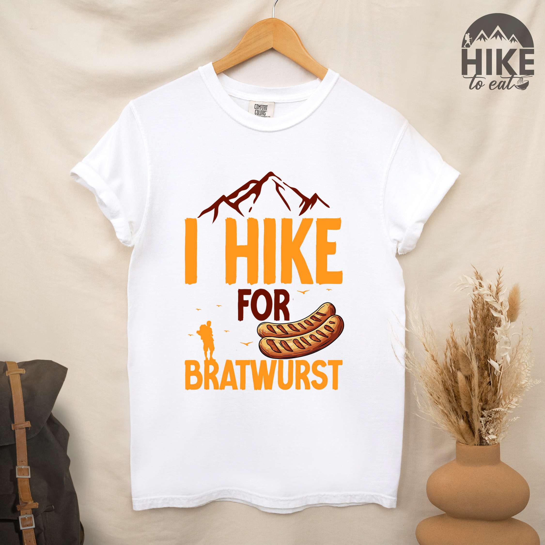 Hanging white "I hike for bratwurst" colored vintage tee with bold typography, illustrated bratwurst, mountains, birds, and backpackers for street dog lovers.