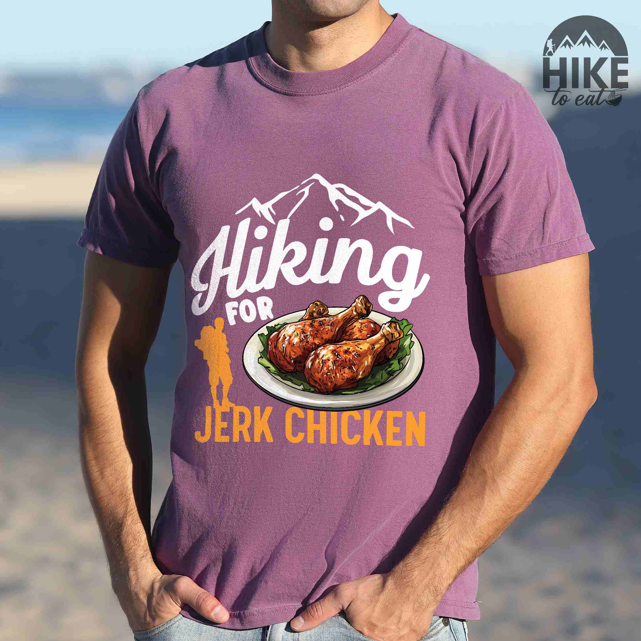 Man wearing berry "hiking for jerk chicken" colored vintage tee with bold typography, illustrated jerk chicken on a plate, mountains, birds, and backpackers for bbq chicken lovers.