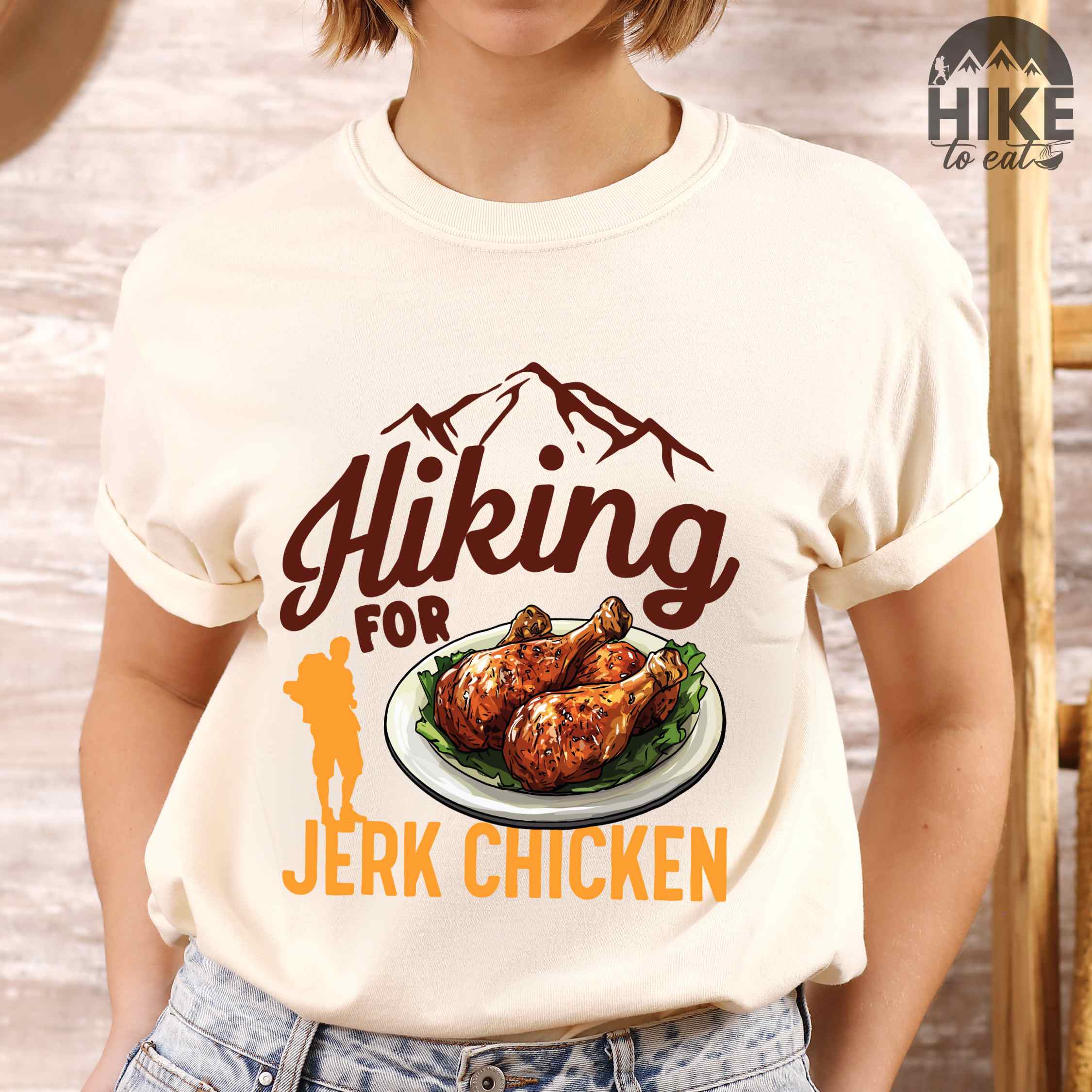 Woman wearing ivory "hiking for jerk chicken" colored vintage tee with bold typography, illustrated jerk chicken on a plate, mountains, birds, and backpackers for bbq chicken lovers.