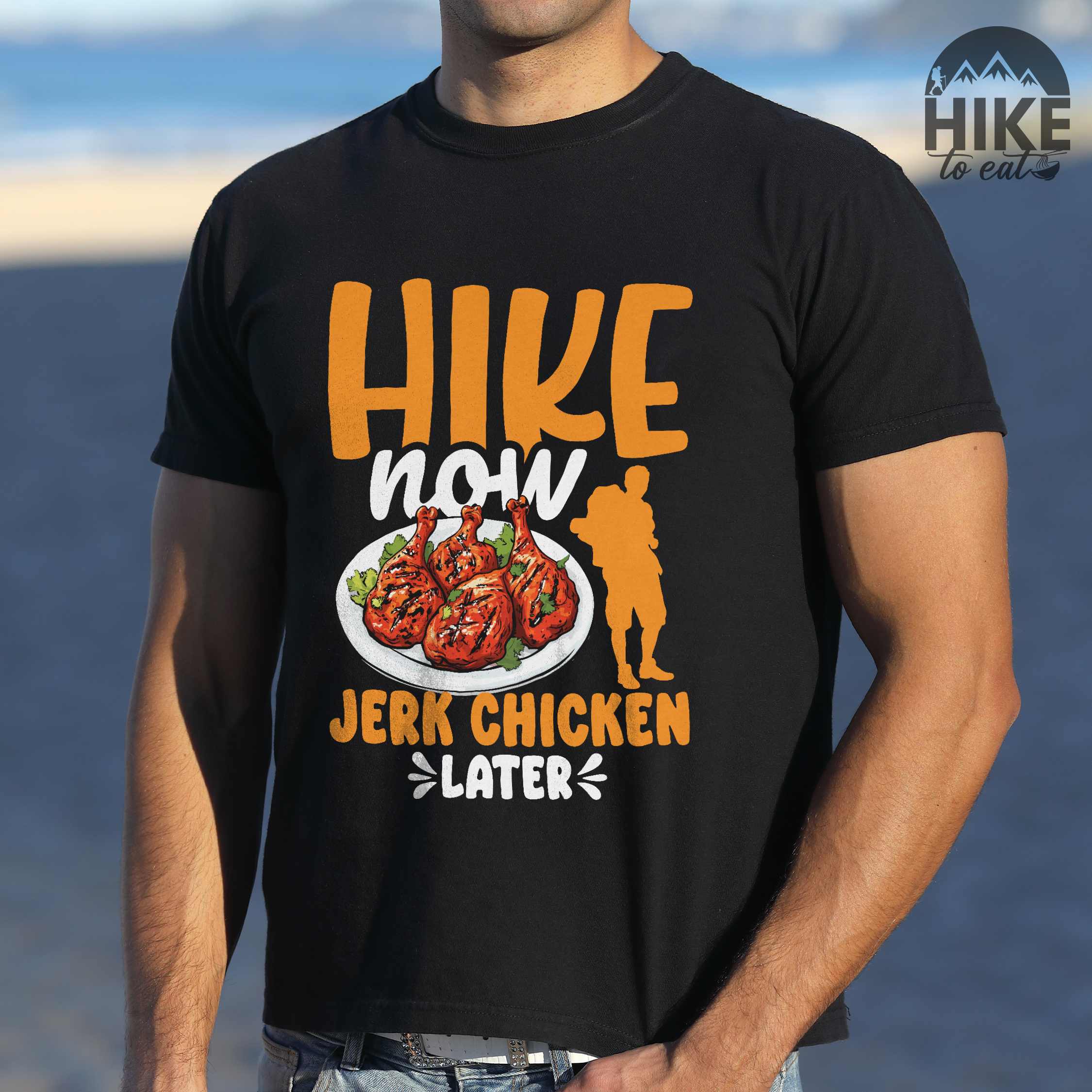 Man wearing black "hike now jerk chicken later" colored vintage tee with bold typography, illustrated jerk chicken on a plate, mountains, birds, and backpackers for bbq chicken lovers.