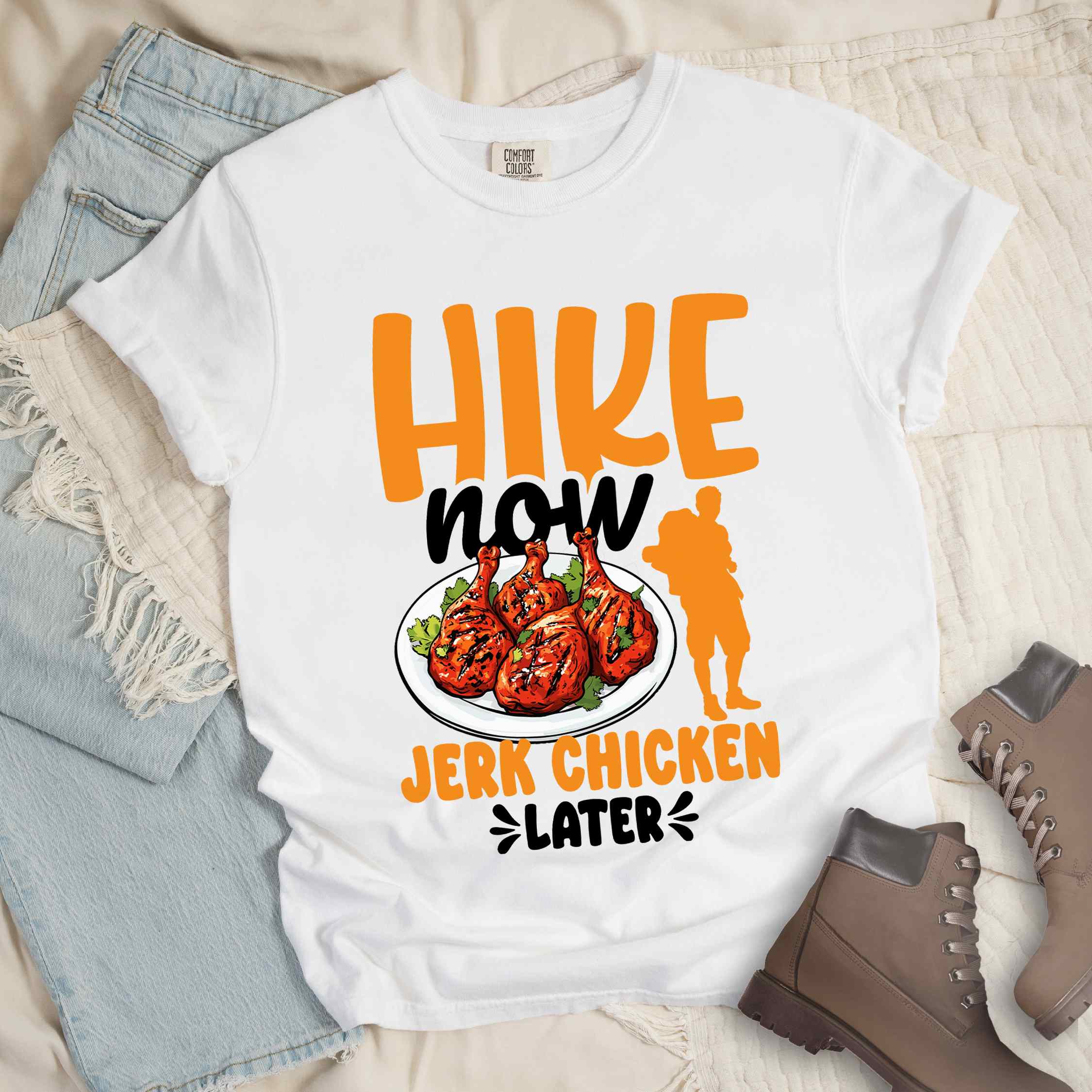 White "hike now jerk chicken later" colored vintage tee with bold typography, illustrated jerk chicken on a plate, mountains, birds, and backpackers for bbq chicken lovers.
