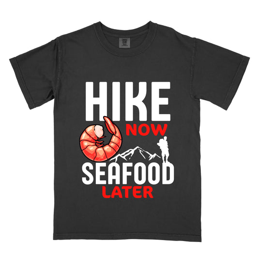 Black t-shirt with white and red text reading "HIKE NOW SEAFOOD LATER". "HIKE" appears in large white letters at the top, "NOW" in red below it, "SEAFOOD" in white in the middle, and "LATER" in red at the bottom. Features an illustration of a red shrimp to the left of "NOW" and white mountain outlines with a white silhouette of a hiker positioned at the right edge of the mountains above the word "SEAFOOD".