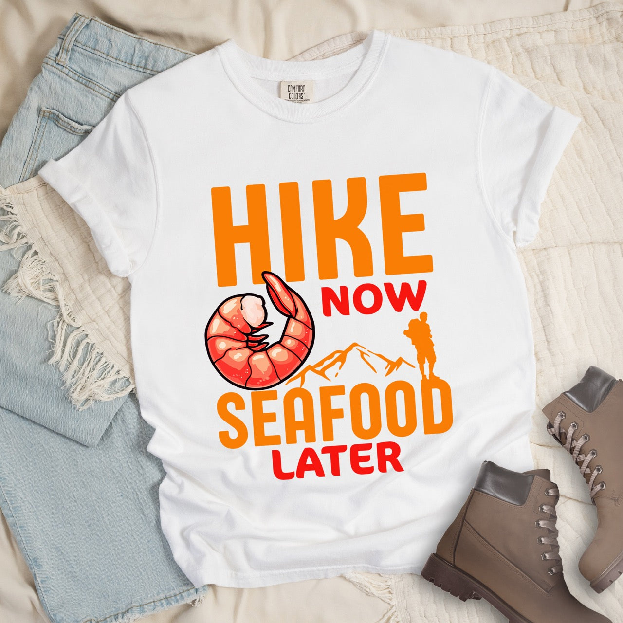 Funny hiking motivation shirt for seafood lovers featuring bold NOW LATER typography, realistic shrimp graphic, mountain landscape illustration, and hiker silhouette on premium outdoor adventure tee perfect gift for trail enthusiasts who love fresh ocean food in white, with jeans and boots