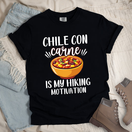 Black t-shirt with white text reading "CHILE CON carne IS MY HIKING MOTIVATION." The word "carne" appears in stylized script. In the center is a colorful illustration of a bowl of chili con carne showing beans, meat, corn, and vegetables in an orange bowl. The shirt features a classic crew neck and casual rolled sleeves, styled alongside light wash denim and brown hiking boots.