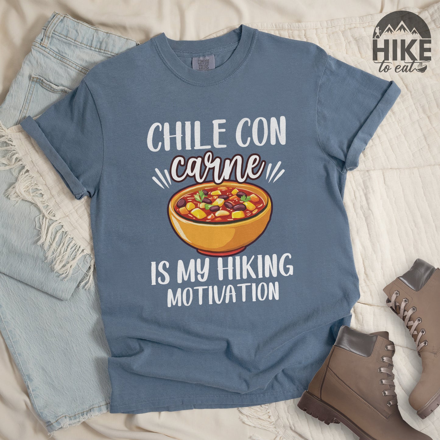 Blue Jean (Denim Blue) t-shirt with white text reading "CHILE CON carne IS MY HIKING MOTIVATION." The word "carne" appears in stylized script. In the center is a colorful illustration of a bowl of chili con carne showing beans, meat, corn, and vegetables in an orange bowl. The shirt features a classic crew neck and casual rolled sleeves, styled alongside light wash denim and brown hiking boots. 