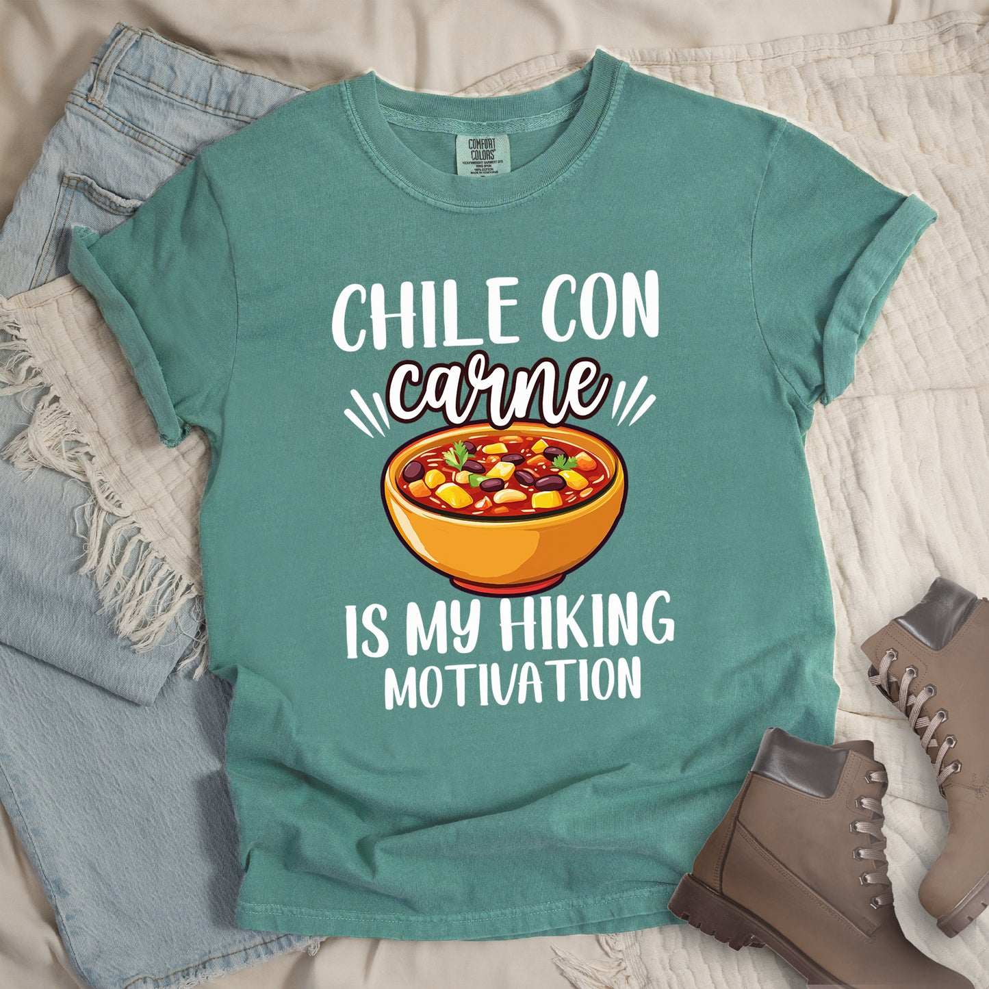 Light Green t-shirt with white text reading "CHILE CON carne IS MY HIKING MOTIVATION." The word "carne" appears in stylized script. In the center is a colorful illustration of a bowl of chili con carne showing beans, meat, corn, and vegetables in an orange bowl.  The shirt features a classic crew neck and casual rolled sleeves, styled alongside light wash denim and brown hiking boots.