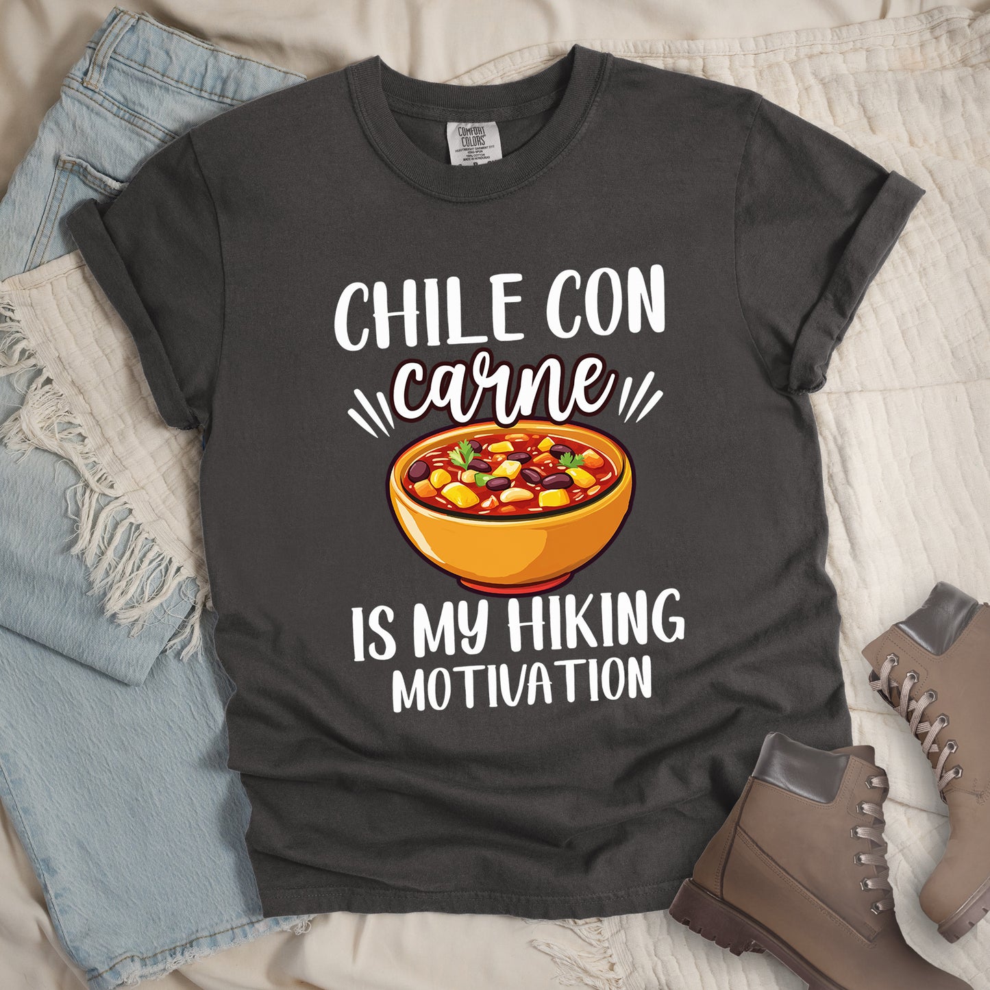 Pepper (Dark Gray) t-shirt with white text reading "CHILE CON carne IS MY HIKING MOTIVATION." The word "carne" appears in stylized script. In the center is a colorful illustration of a bowl of chili con carne showing beans, meat, corn, and vegetables in an orange bowl.  The shirt features a classic crew neck and casual rolled sleeves, styled alongside light wash denim and brown hiking boots.