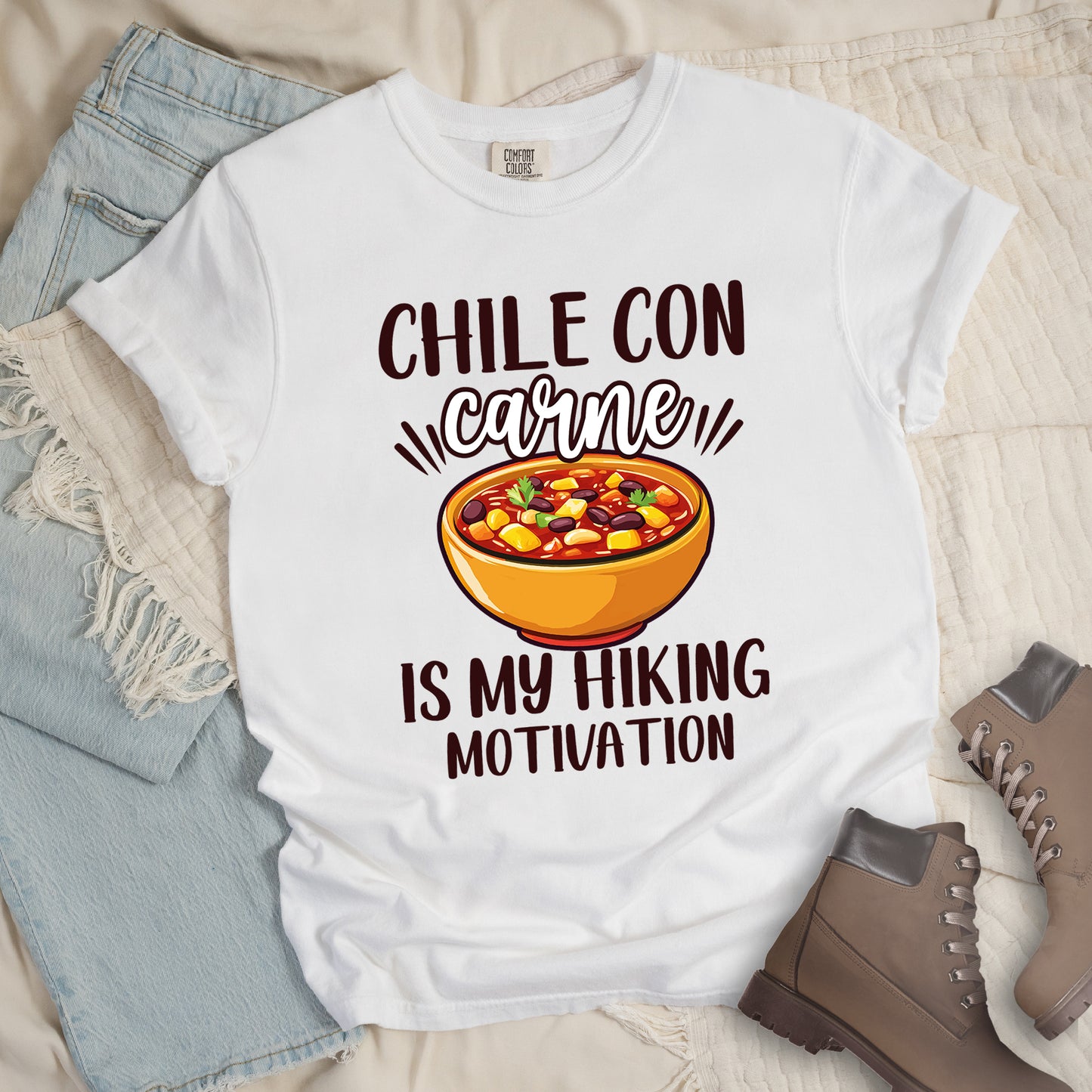 White t-shirt with black text reading "CHILE CON carne IS MY HIKING MOTIVATION." The word "carne" appears in stylized script. In the center is a colorful illustration of a bowl of chili con carne showing beans, meat, corn, and vegetables in an orange bowl. The shirt features a classic crew neck and casual rolled sleeves, styled alongside light wash denim and brown hiking boots.